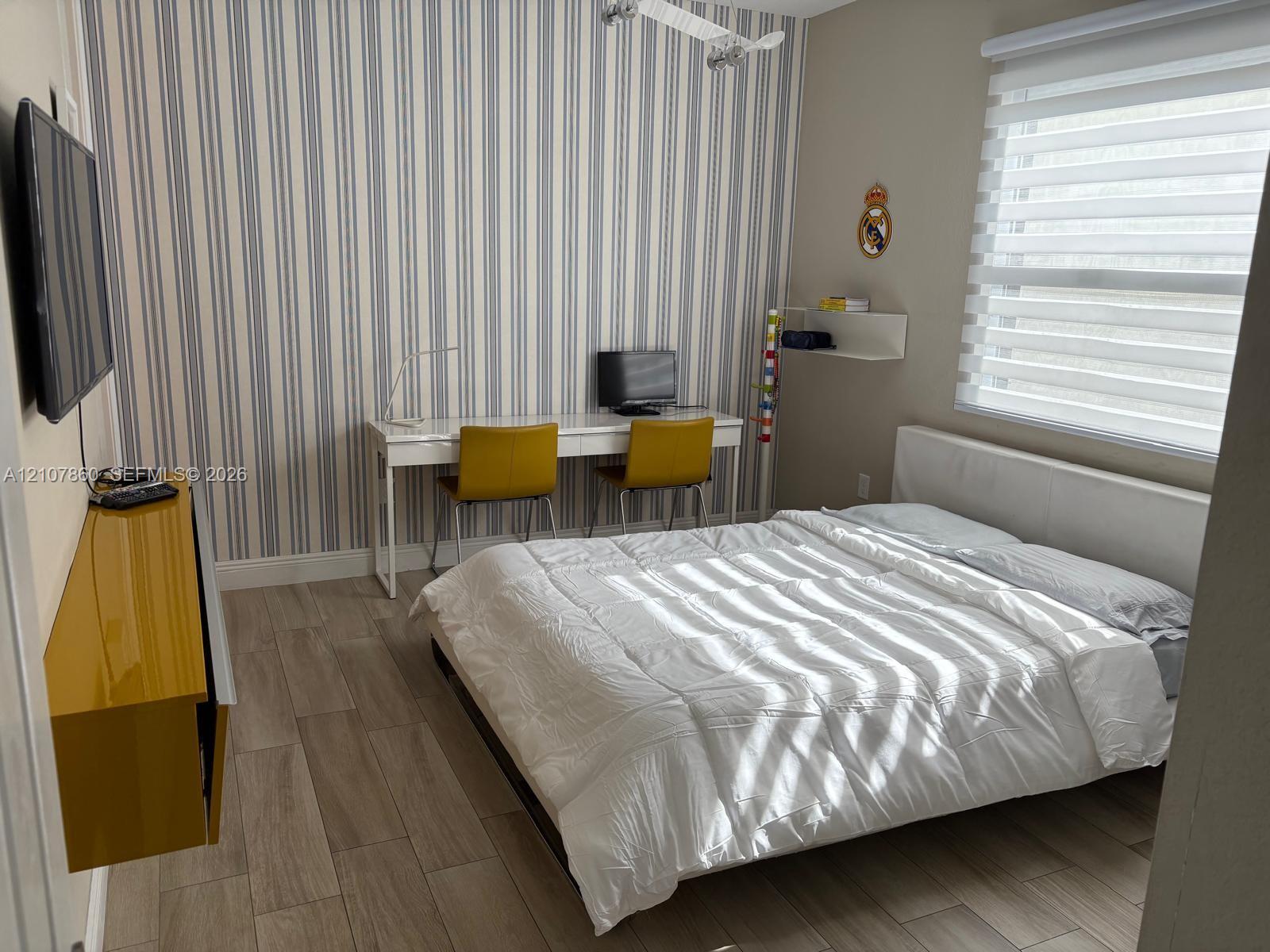 Wood-finish flooring, vertical-striped accent wall, built-in mustard-tone wall cabinetry, and light-filtering horizontal window shades