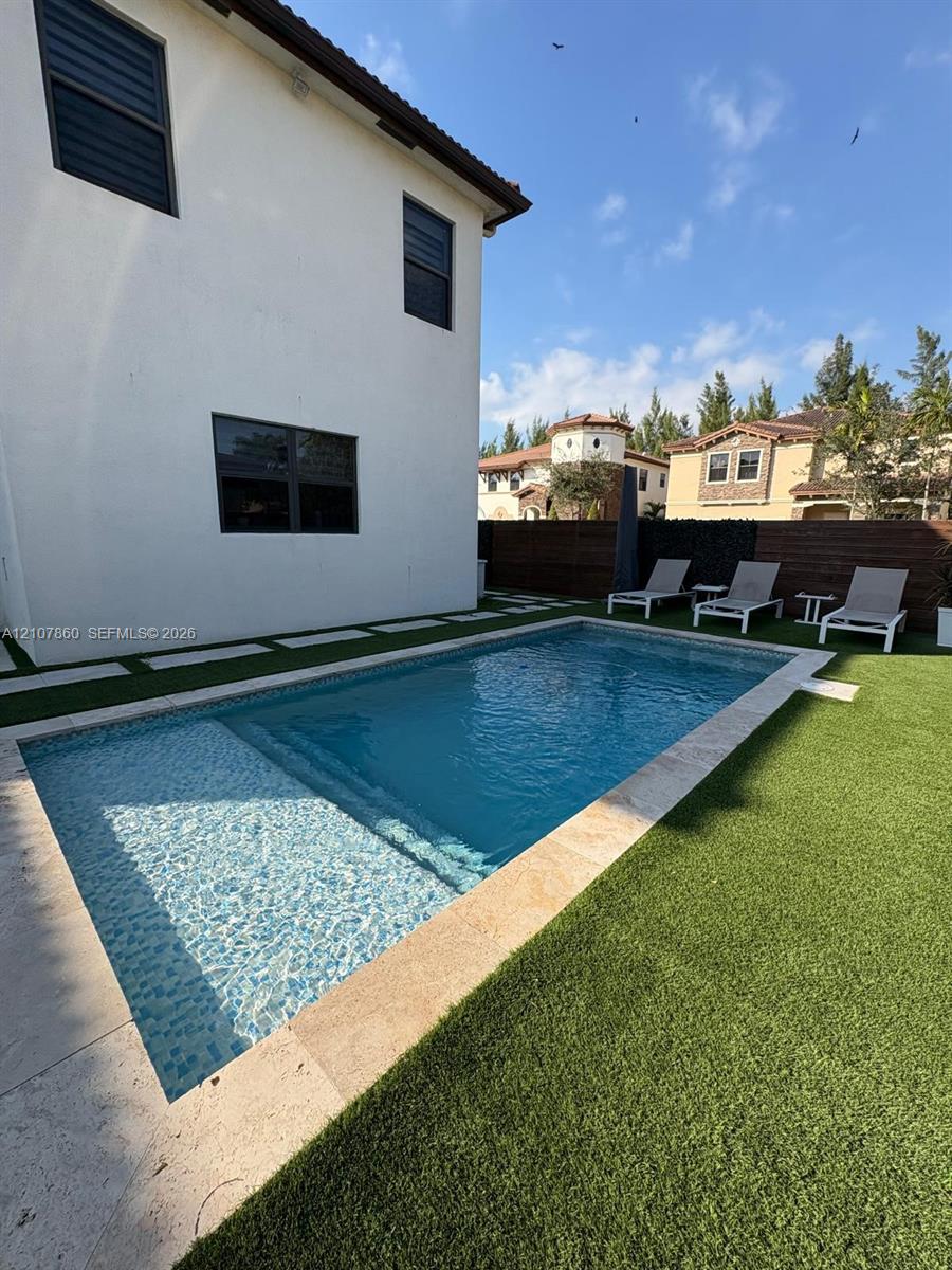 Rectangular swimming pool featuring a tanning ledge and natural stone coping, surrounded by synthetic turf and oversized concrete pavers, enclosed by a horizontal wood fence
