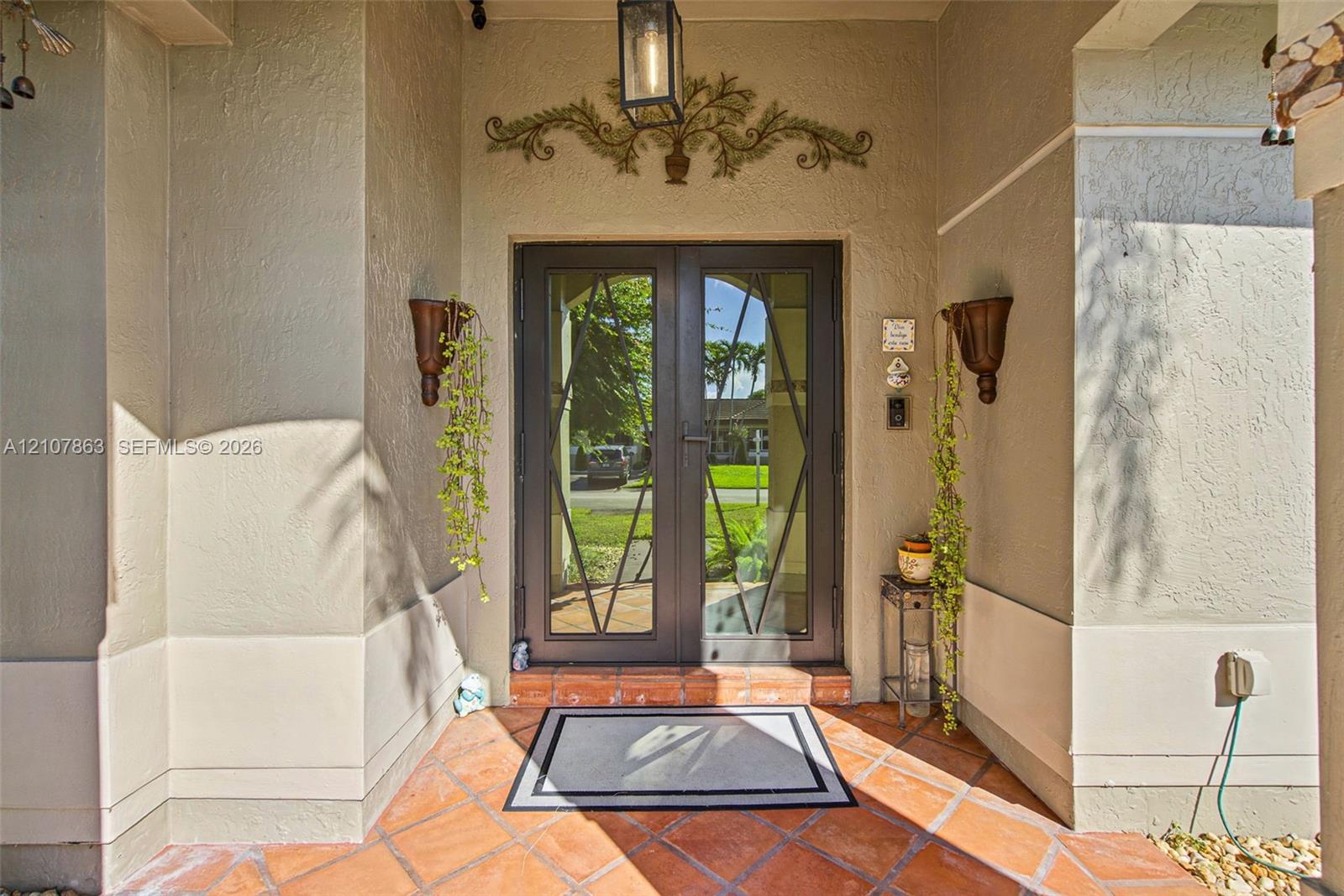 Covered entryway featuring double doors with geometric glass inserts, terracotta-toned tile flooring, stucco exterior walls, and a decorative metal wall accent