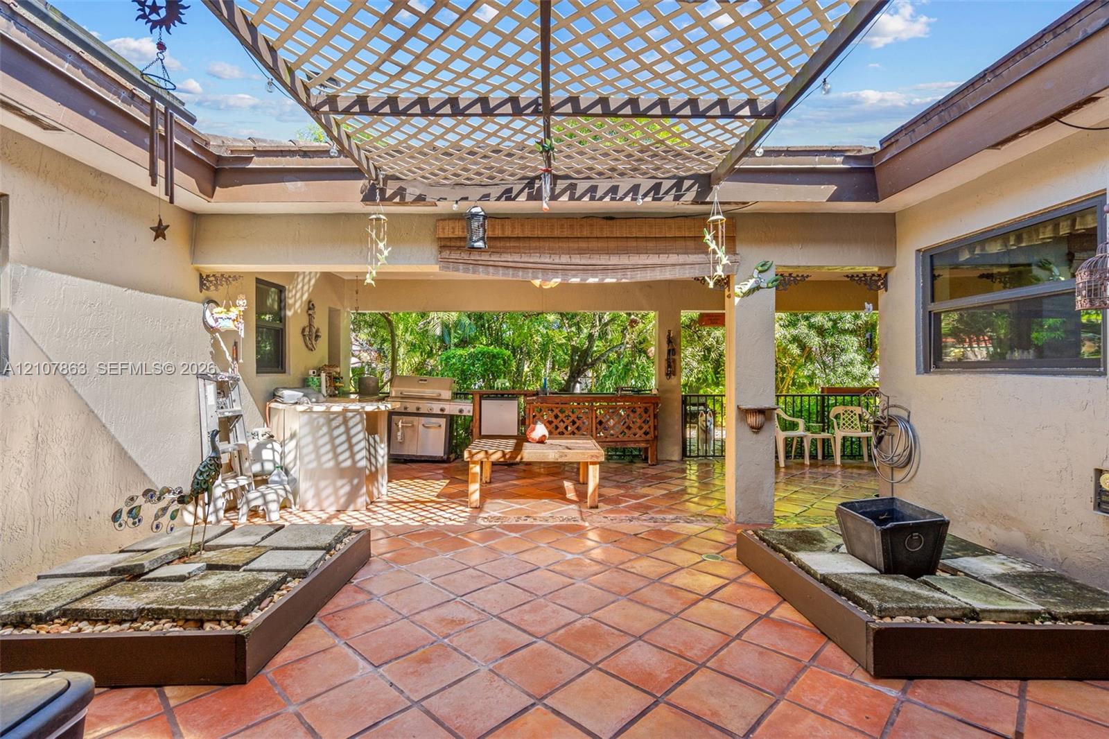 Covered outdoor patio featuring tile flooring, built-in outdoor kitchen, lattice overhead structure, and dual raised stone planters
