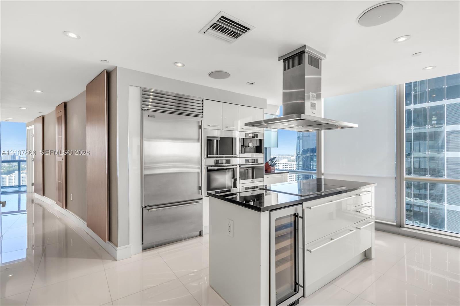Modern kitchen featuring high-gloss cabinetry, stainless steel professional-grade appliances, a central island with a built-in wine refrigerator, a stone countertop, and floor-to-ceiling windows