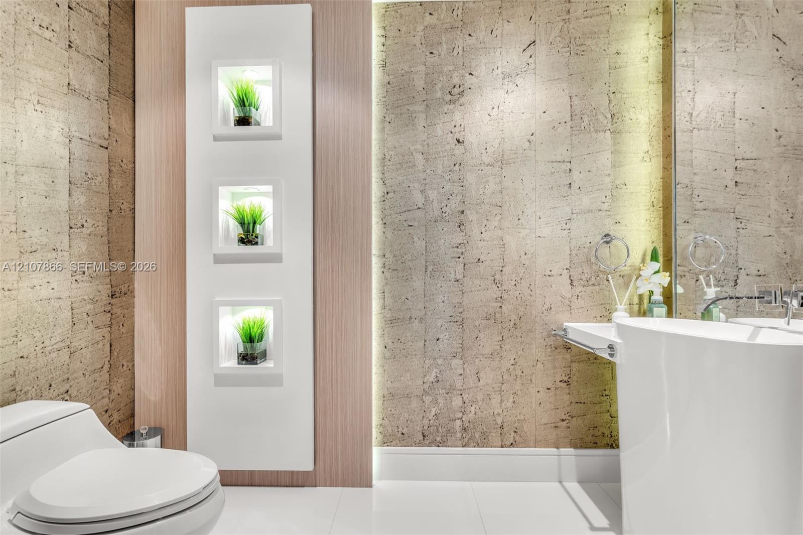 Textured stone-finish wall coverings, vertical built-in recessed shelving with integrated lighting, wood-finish paneling, white pedestal sink, and polished wall-mounted fixtures