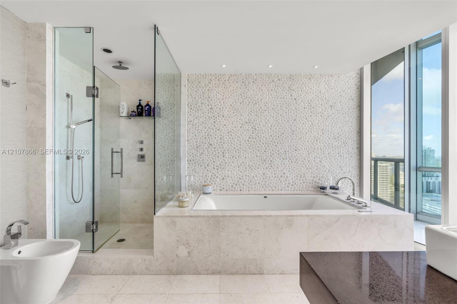 Bathroom featuring a built-in soaking tub, glass-enclosed walk-in shower with a rainfall showerhead, stone-tile flooring, textured stone-accent wall, and floor-to-ceiling windows