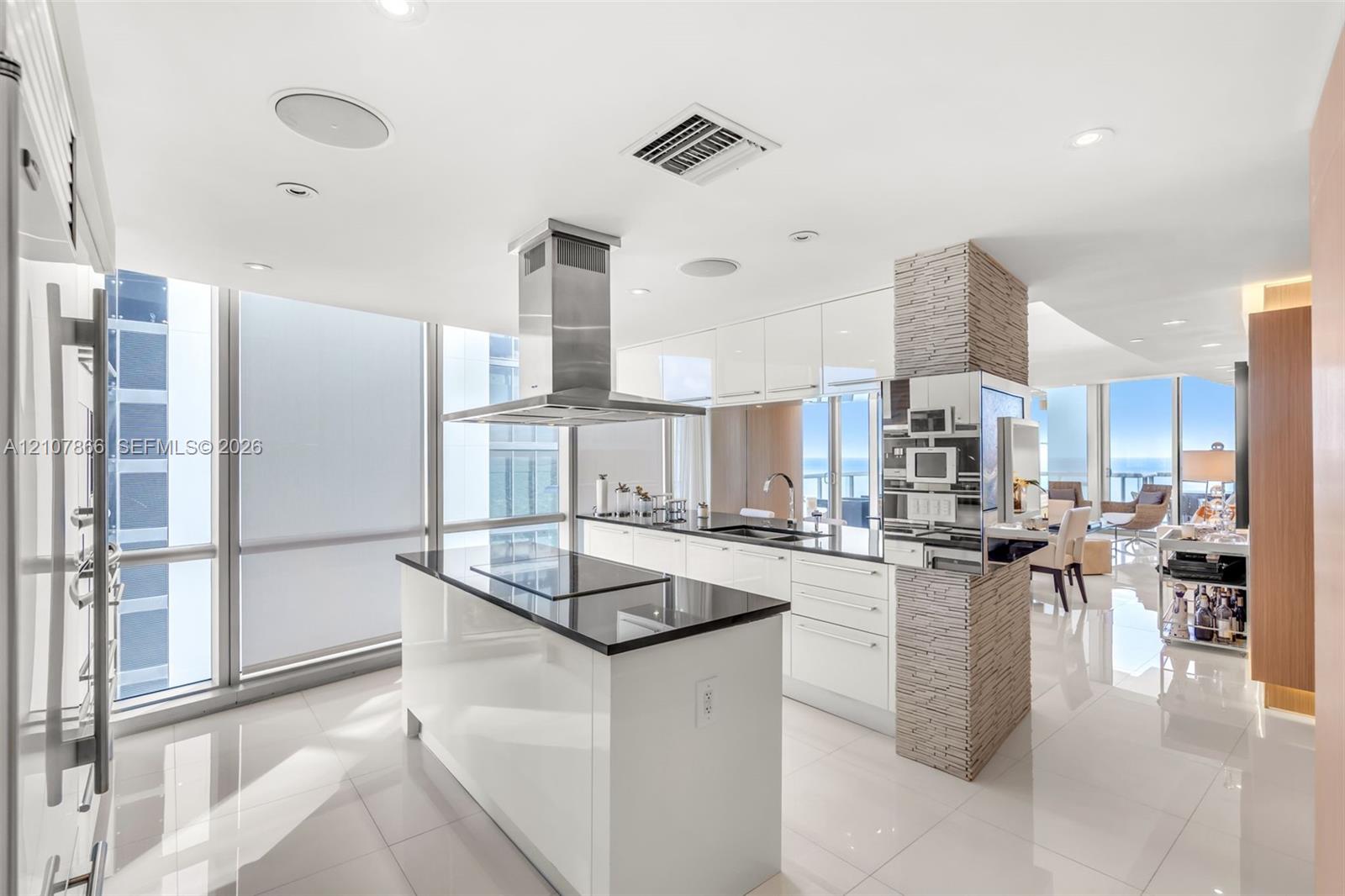 Contemporary kitchen featuring a center island with a sleek cooktop, stainless steel chimney-style range hood, high-gloss cabinetry, floor-to-ceiling windows, and a decorative stone-clad pillar