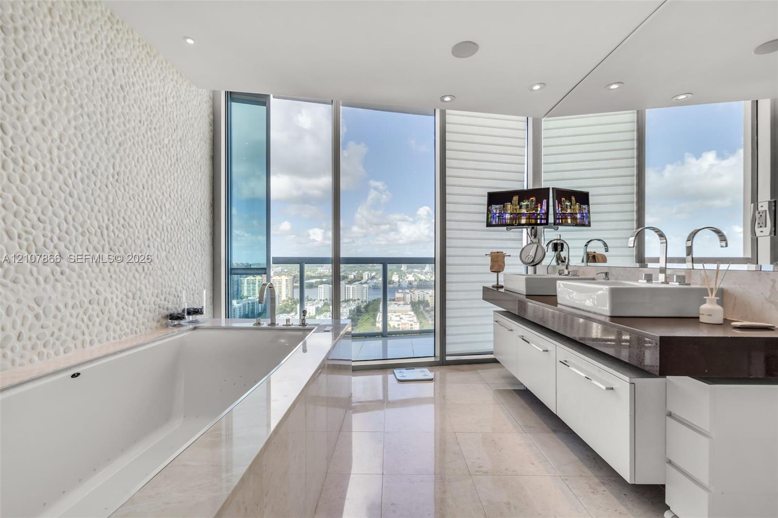 Bathroom featuring floor-to-ceiling windows, a deep soaking tub with a textured stone accent wall, a floating dual-sink vanity with a countertop, and recessed ceiling lighting