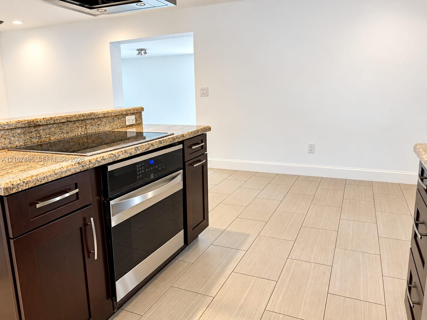 Kitchen featuring dark cabinetry with metallic hardware, stone-finish countertops, an integrated glass cooktop, a stainless steel oven, and light-toned tile flooring