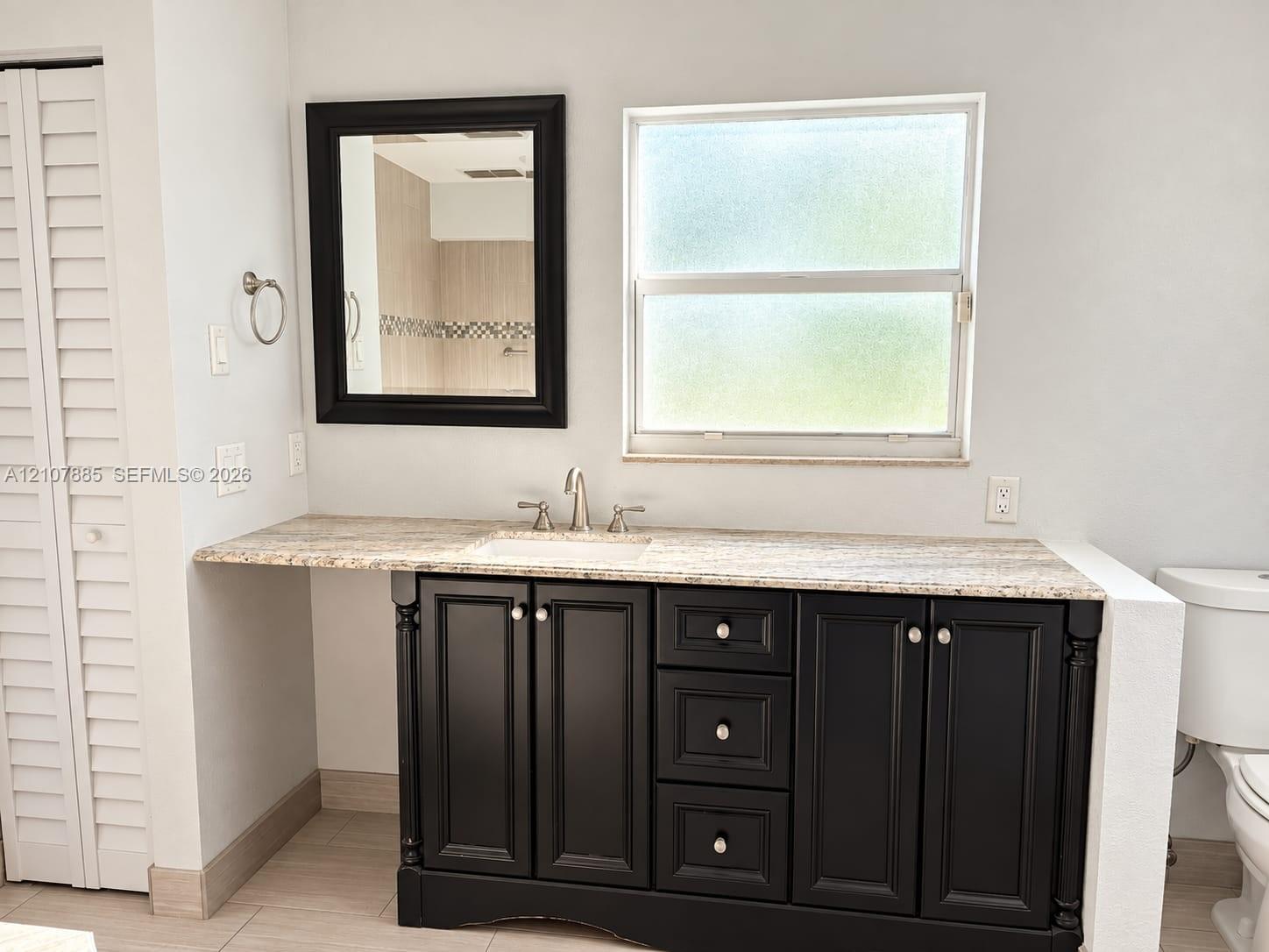 Bathroom vanity featuring dark-finished cabinetry, a light-toned stone countertop, an integrated undermount sink, a framed wall mirror, and dual-handle metal faucet hardware