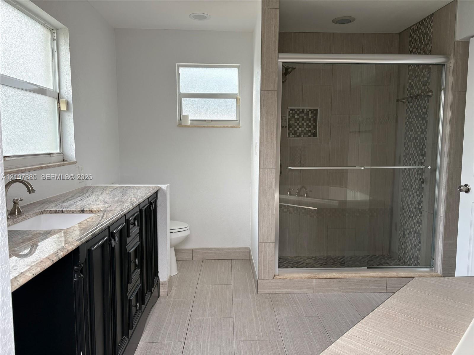Dark cabinetry vanity with stone-finish countertop, undermount sink, walk-in shower with sliding glass doors, mosaic tile accent, and light-tone floor tiling