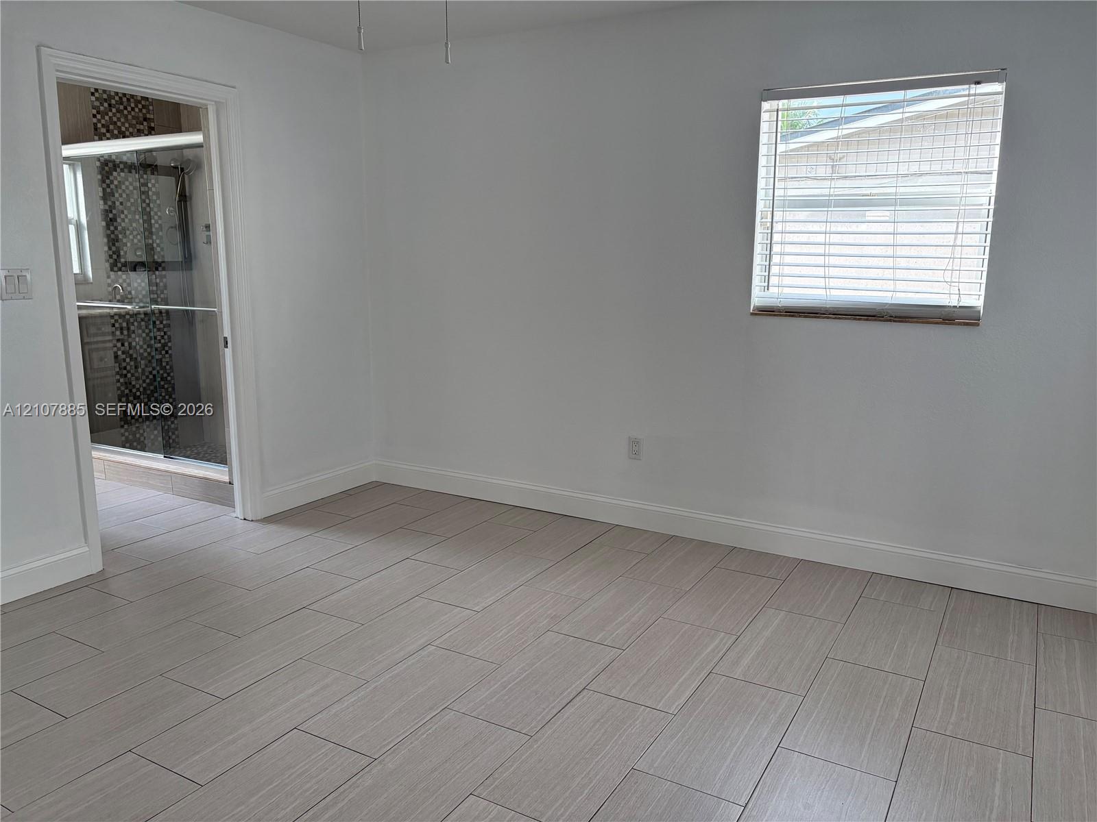 Neutral-toned tile flooring, white wall finish, window with horizontal blinds, and doorway revealing a tiled shower enclosure with decorative mosaic accents