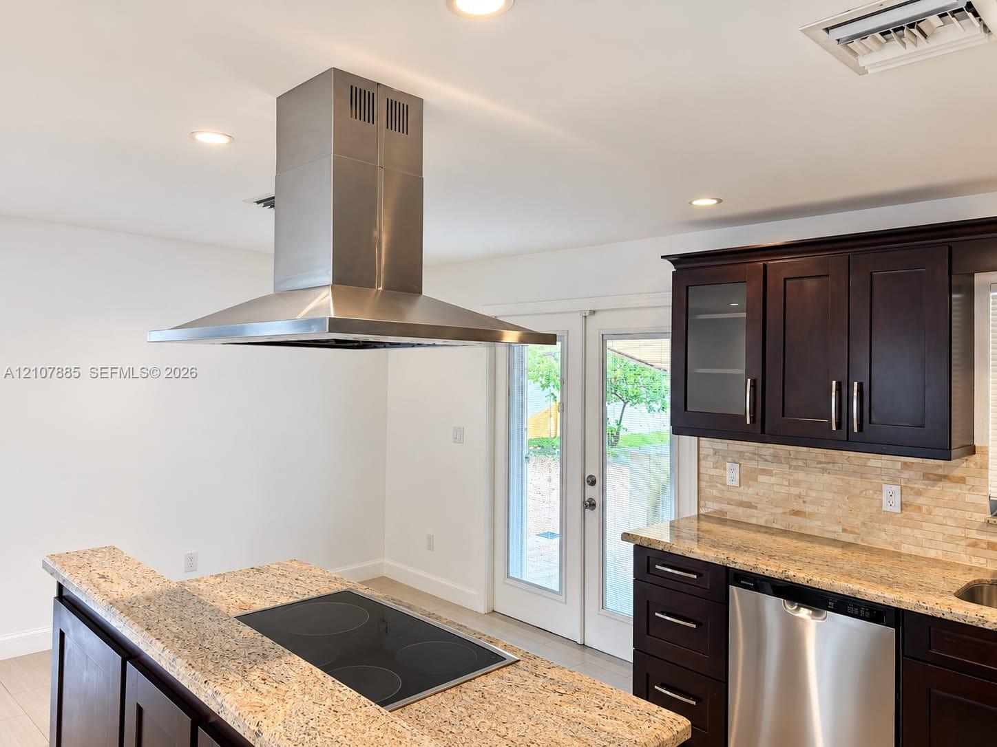 Kitchen featuring dark wood cabinetry, stone countertops, stainless steel appliances, a center island with a built-in cooktop, and a stainless steel vent hood