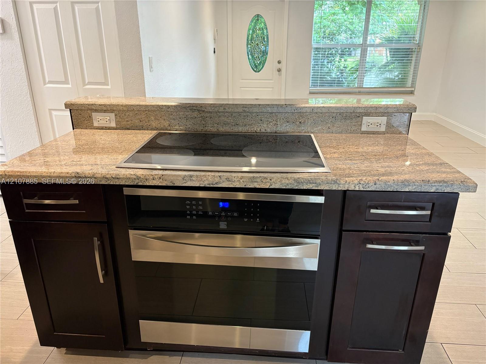 Kitchen island featuring a stone countertop, integrated electric cooktop, stainless steel oven, and dark-toned cabinetry with metal hardware
