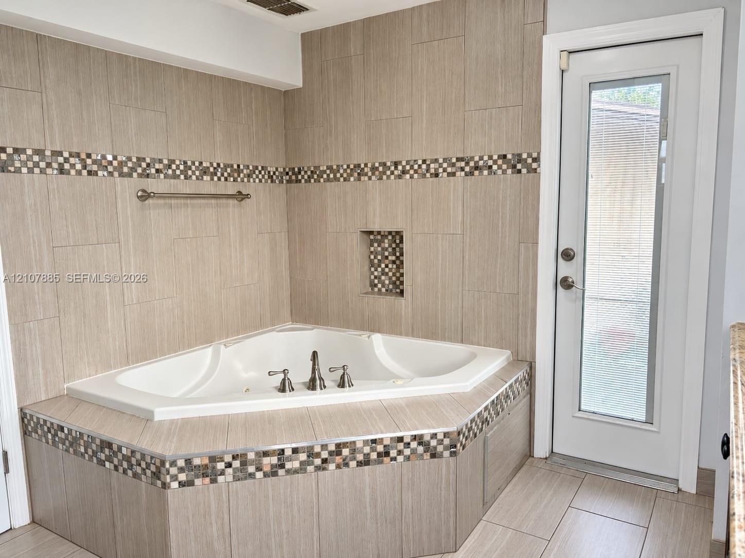 Corner soaking tub featuring a tiled surround, recessed wall niche, multi-colored accent tile detailing, and a glass-paneled exterior door