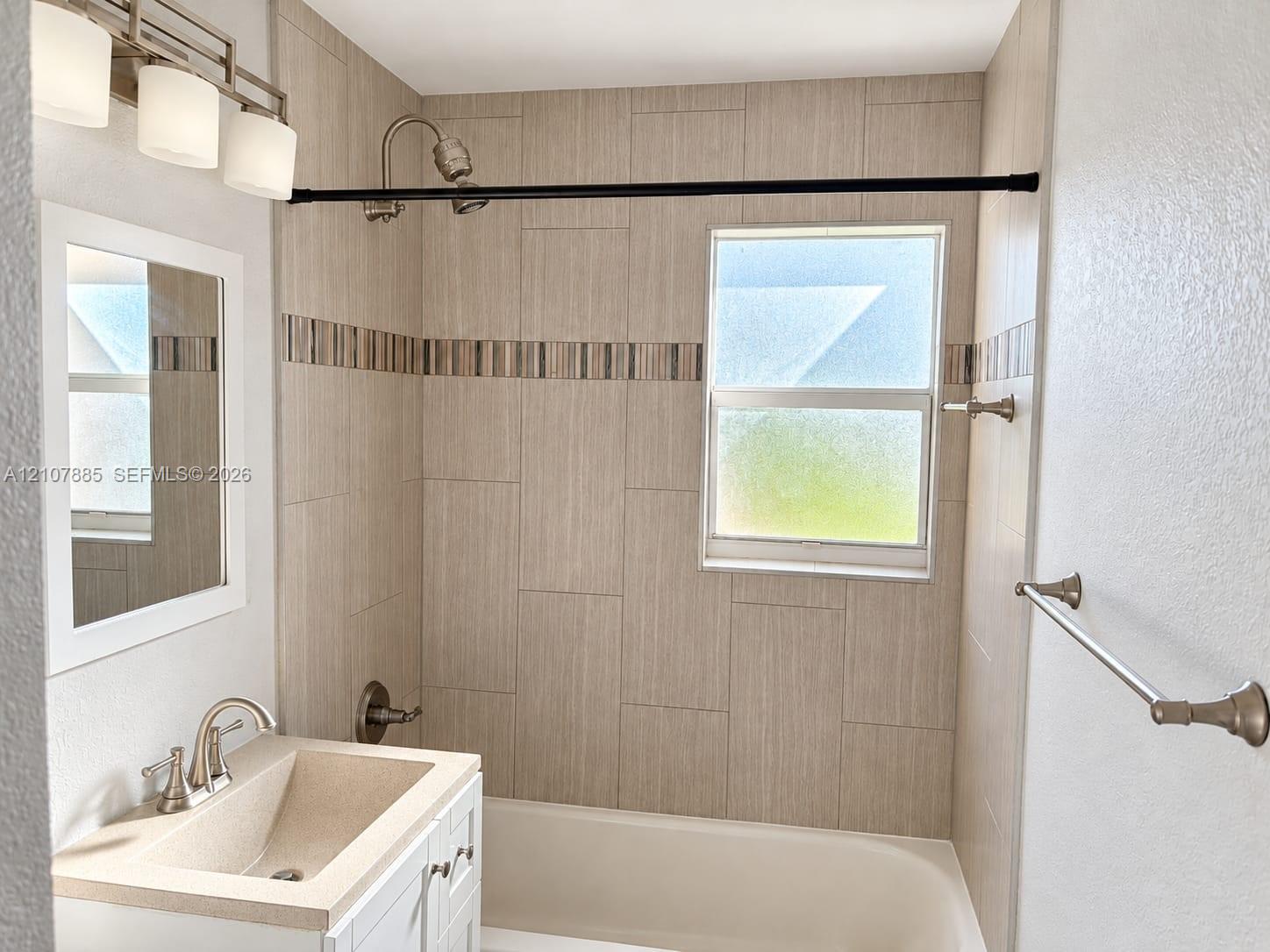 Bathroom featuring a white vanity with an integrated countertop, wall-mounted light fixture, tiled shower surround with decorative accent inlay, and obscured glass window