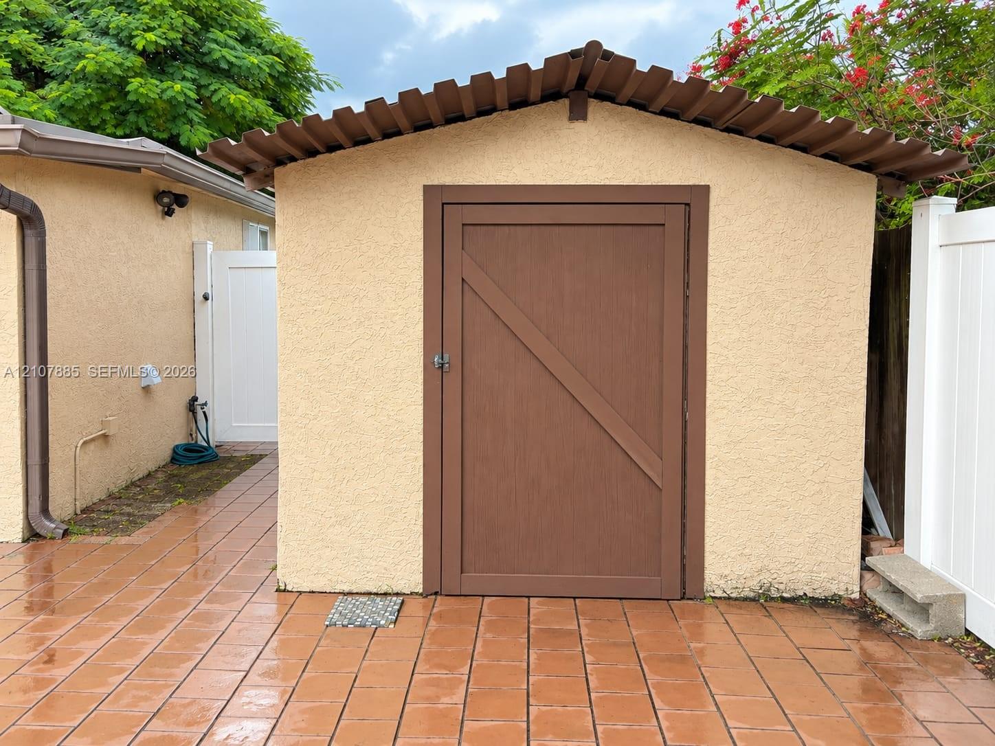 Stucco exterior storage structure featuring a single-panel door with wood-finish trim, a corrugated roof, and matching tiled patio flooring
