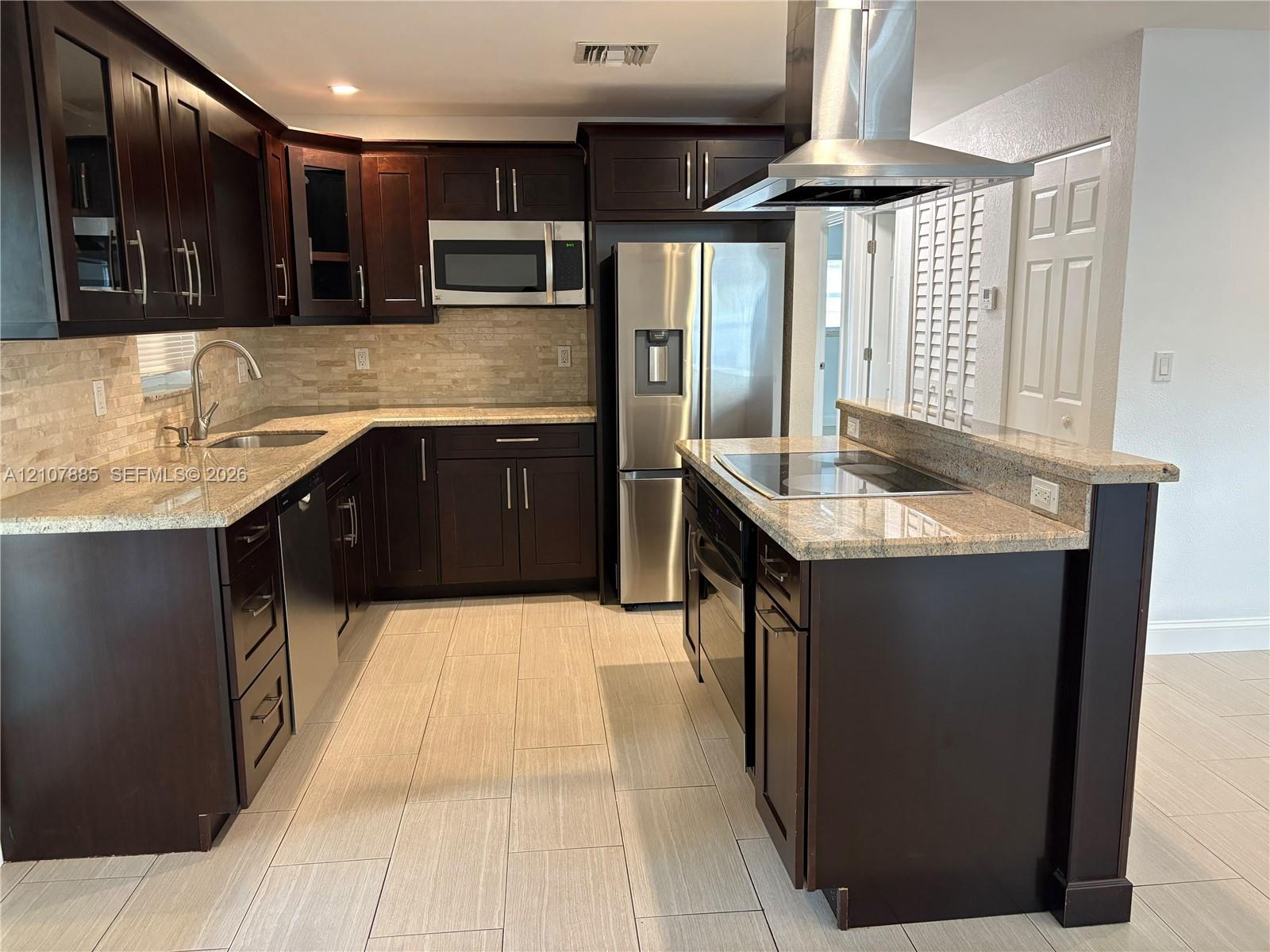 Kitchen featuring dark wood cabinetry, stone countertops, stacked-stone backsplash, stainless steel appliances, and a center island with a built-in cooktop
