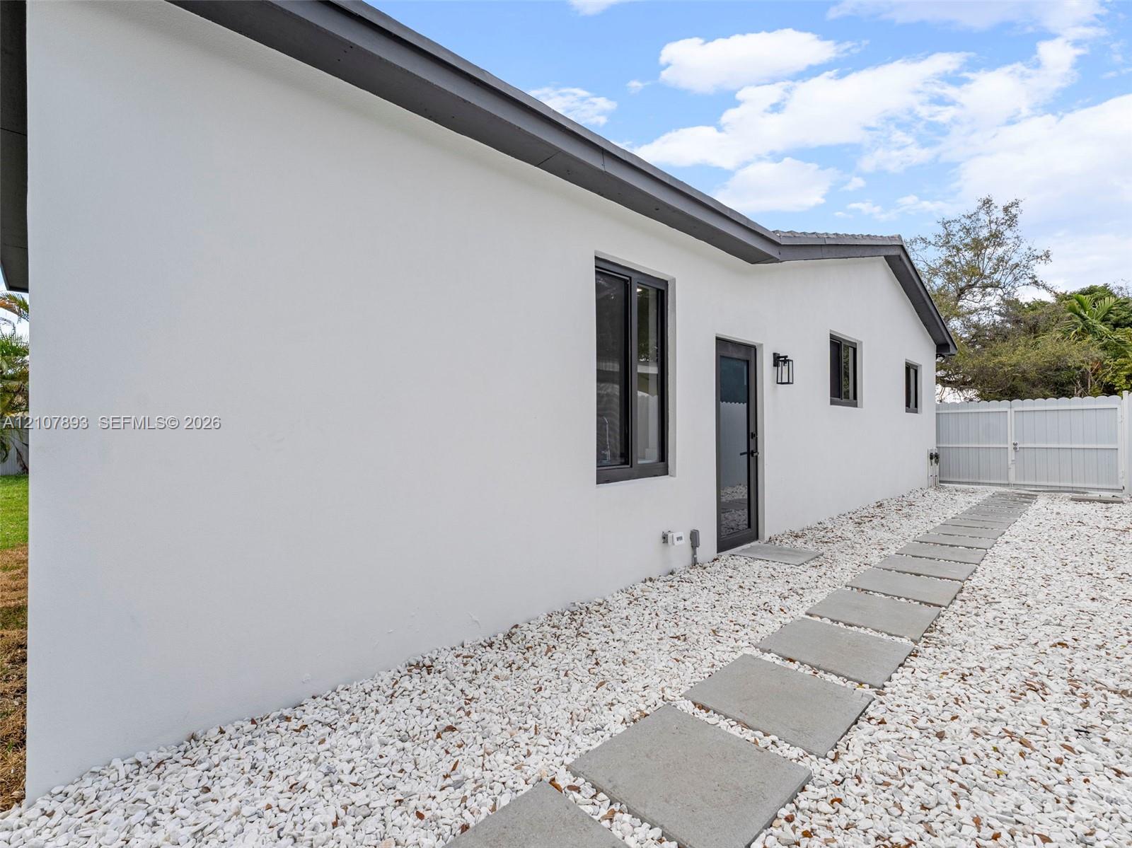 Exterior featuring smooth white stucco finish, dark-trimmed windows, concrete paver walkway, gravel ground cover, and gated side access