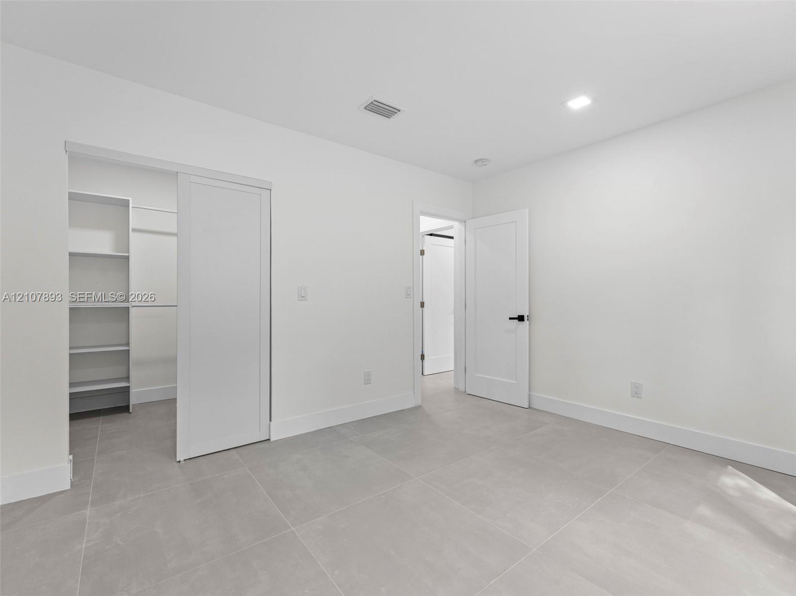 Interior space featuring large-format grey tile flooring, white walls, recessed ceiling lighting, a walk-in closet with built-in shelving, and a solid-core door with matte black hardware