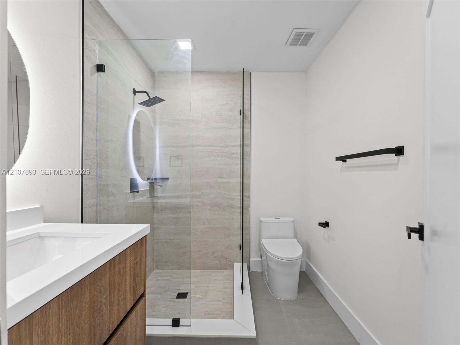 Wood-finish vanity with a rectangular sink, walk-in shower with a frameless glass enclosure, neutral tile surround, matte black rainfall shower head, and an illuminated circular mirror
