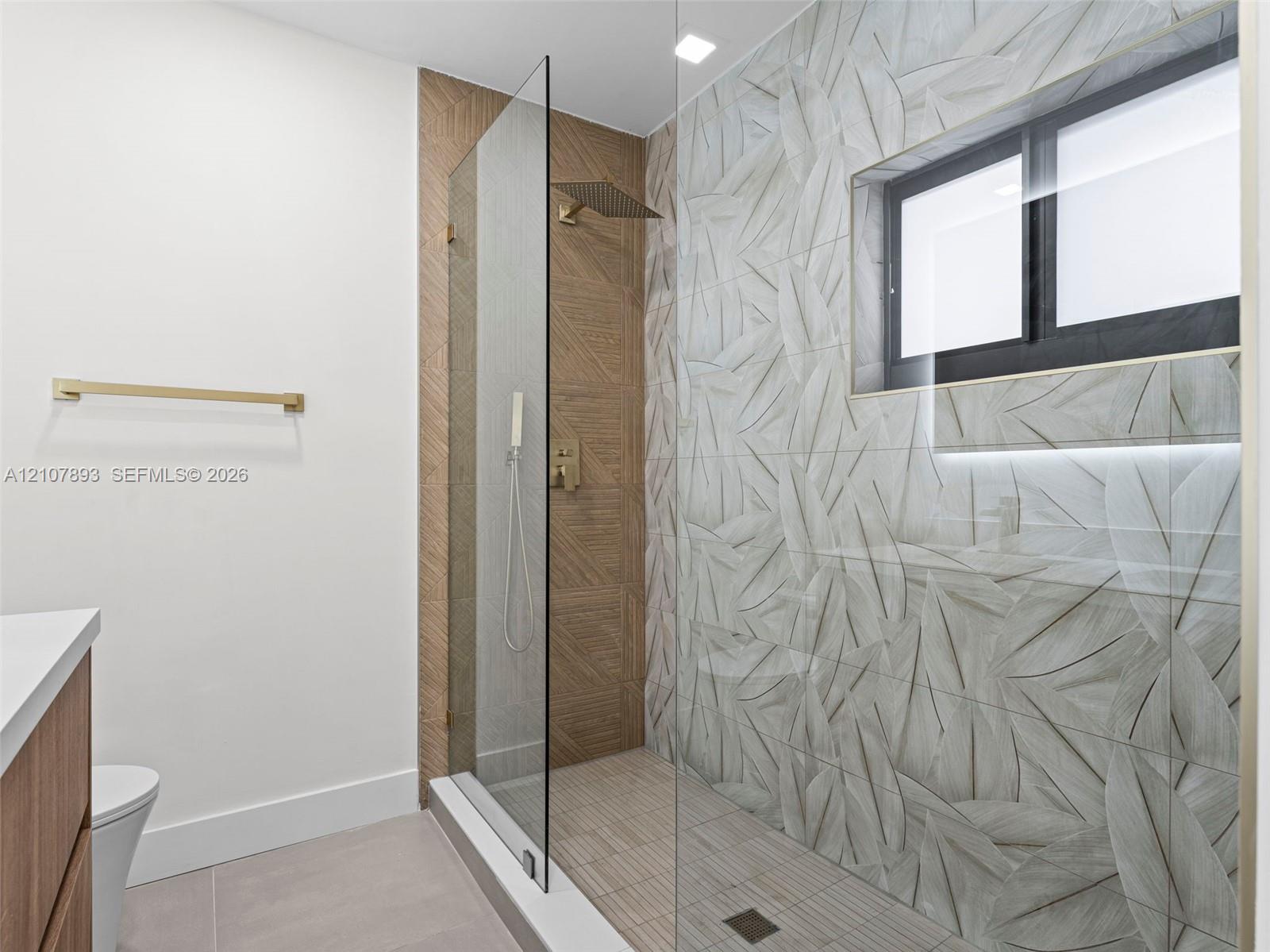 Walk-in shower featuring gold-tone rainfall and handheld showerheads, glass enclosure, leaf-patterned wall tile, and built-in niche lighting