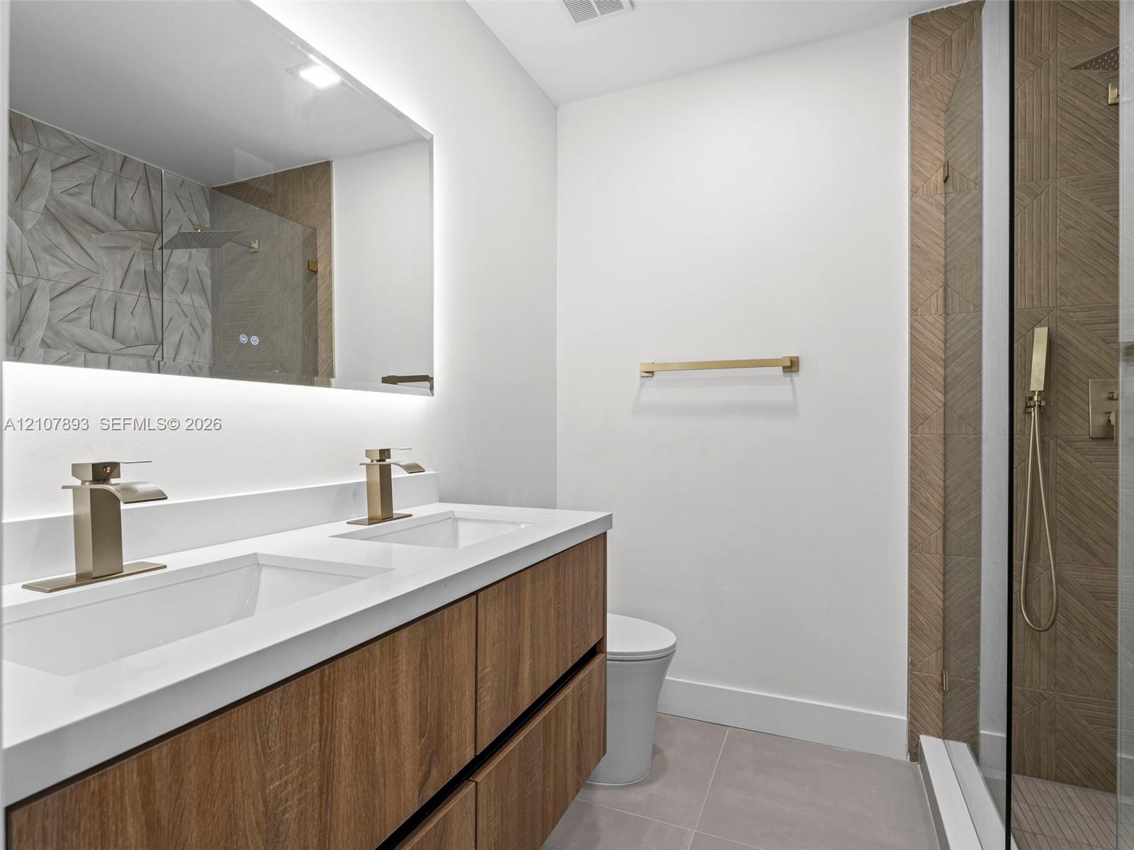 Floating wood-finish double vanity with white countertop, integrated rectangular sinks, brass-toned fixtures, LED-backlit mirror, and glass-enclosed shower with patterned tile surround
