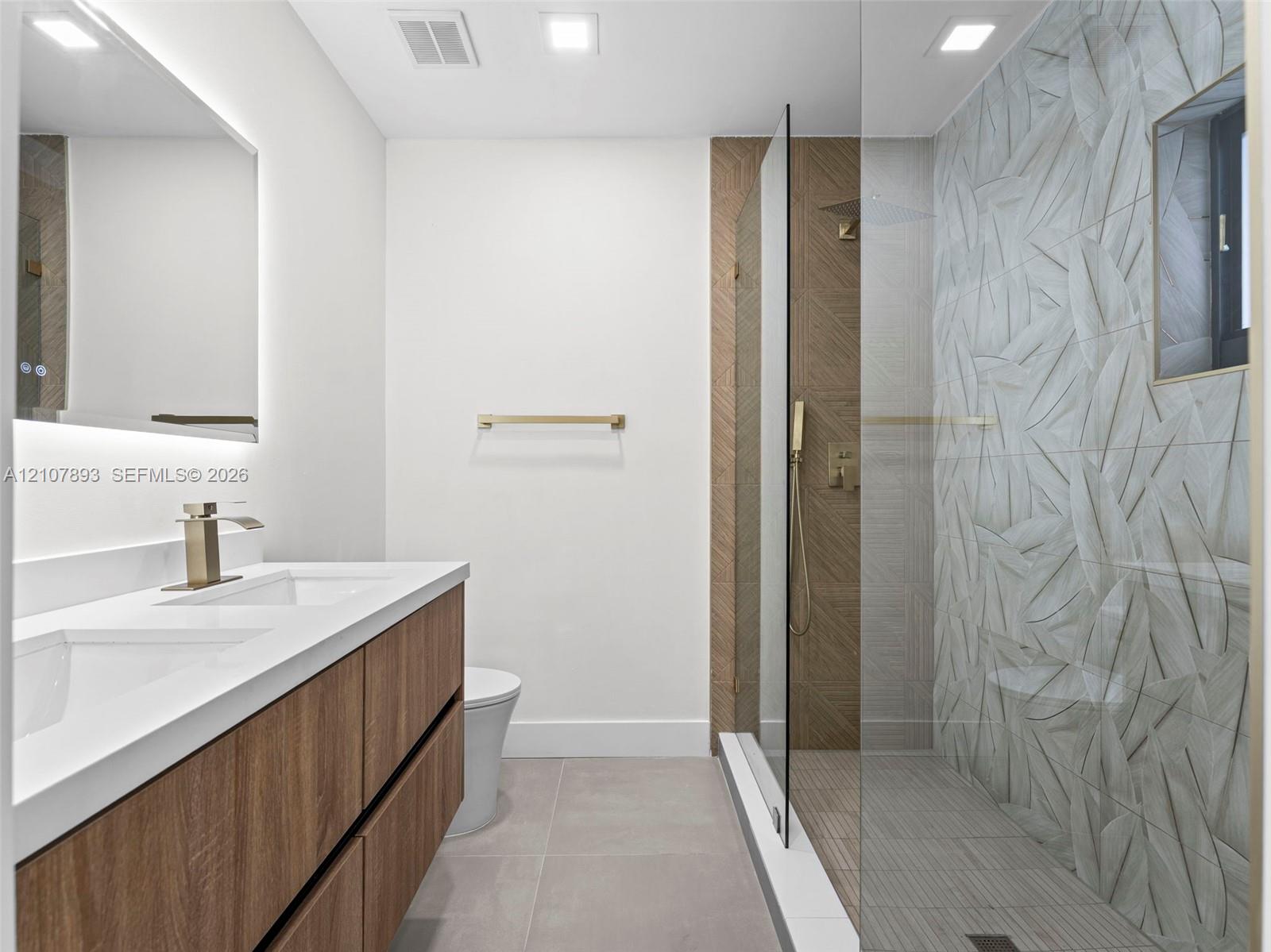 Double vanity with quartz countertop, wood-finish cabinetry, frameless glass-enclosed shower featuring textured leaf-patterned wall tile, and matte gold-tone fixtures
