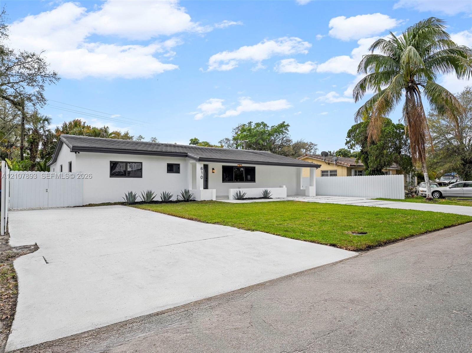 Single-story exterior featuring white stucco finish, dark-toned hip roof, expansive concrete driveway, manicured lawn, and white perimeter fencing