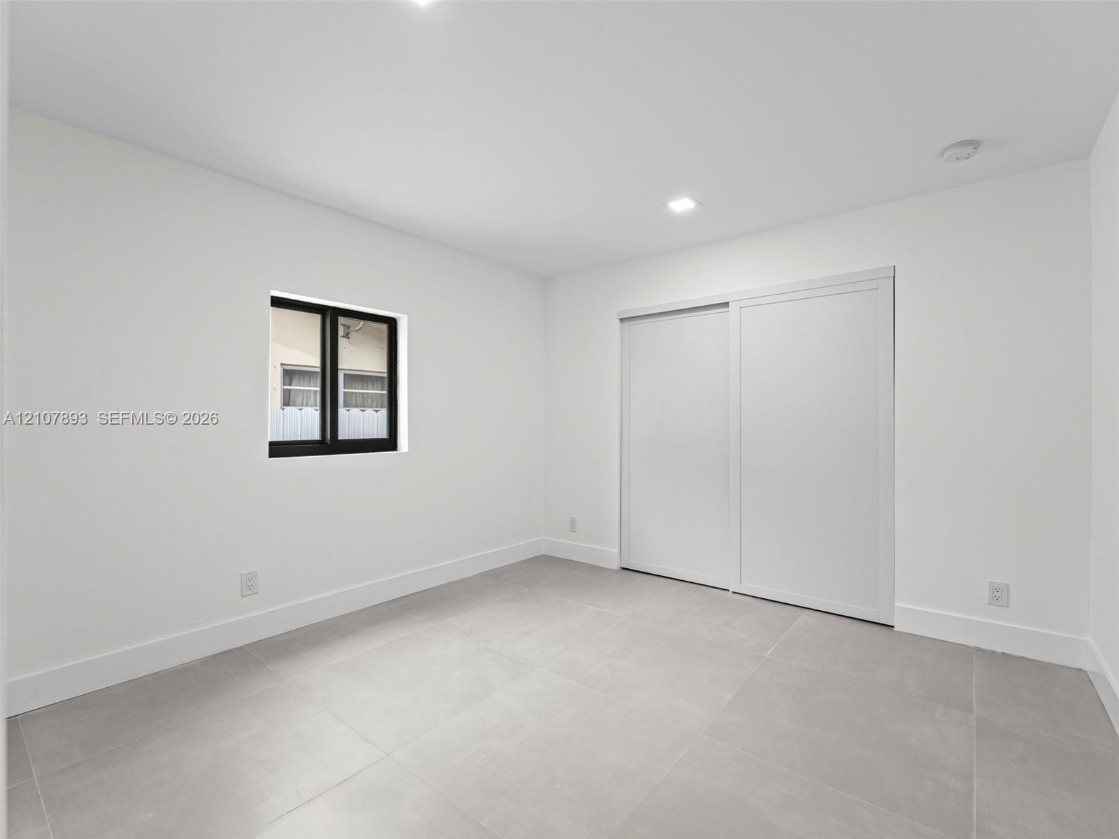 Neutral-toned large-format tile flooring, recessed ceiling lighting, sliding-panel closet doors, and a window with a black frame