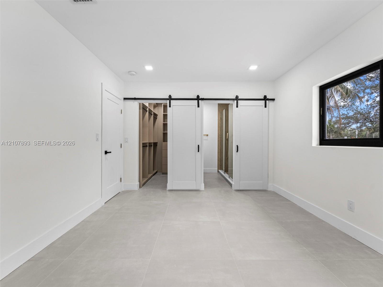 Large-format tile flooring, dual sliding barn-style doors with matte black hardware, built-in closet storage system, and recessed lighting