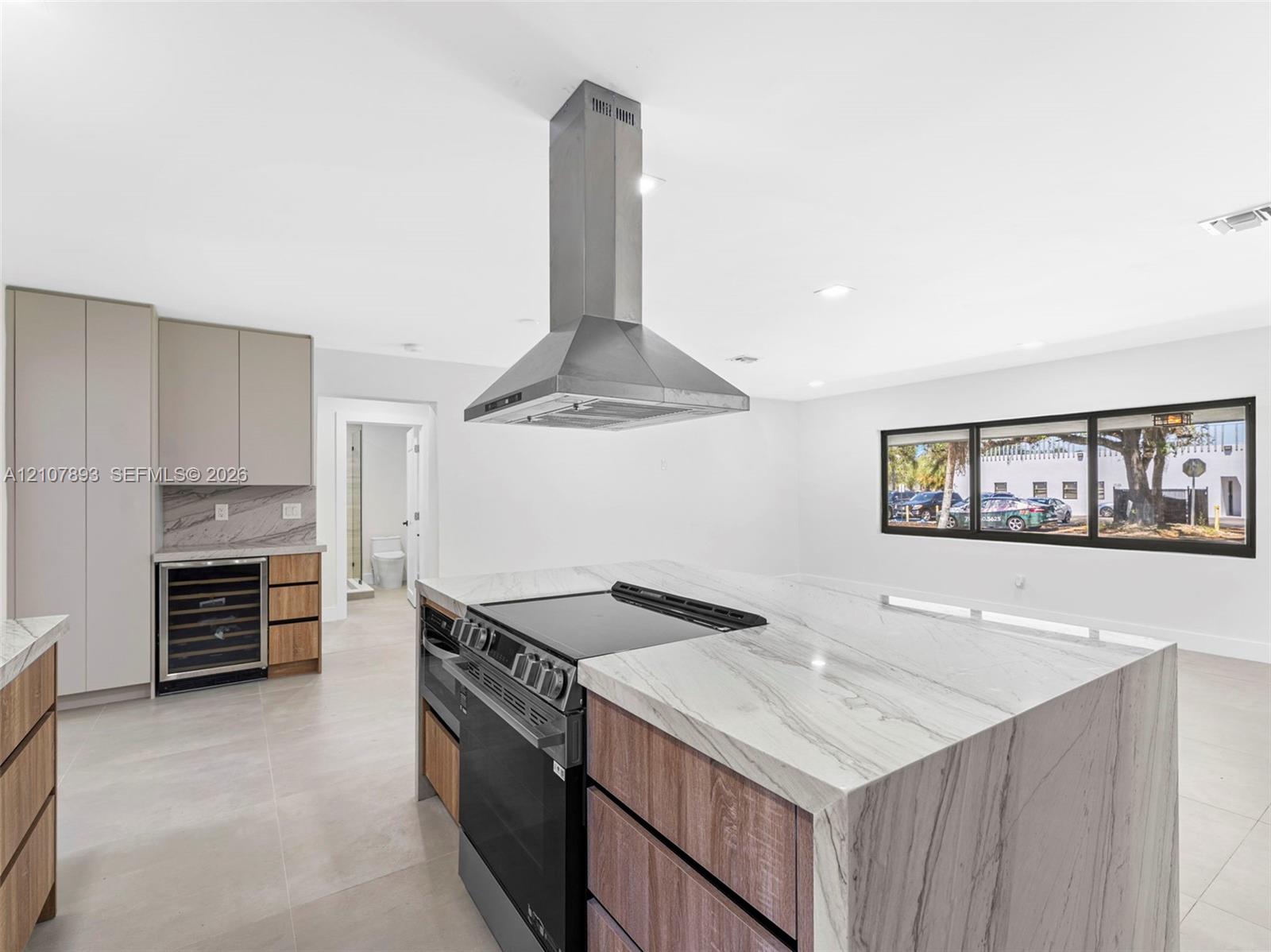 Kitchen island featuring waterfall-edge stone countertop, stainless steel range, built-in wine refrigerator, custom wood-finish cabinetry, and stainless steel chimney hood