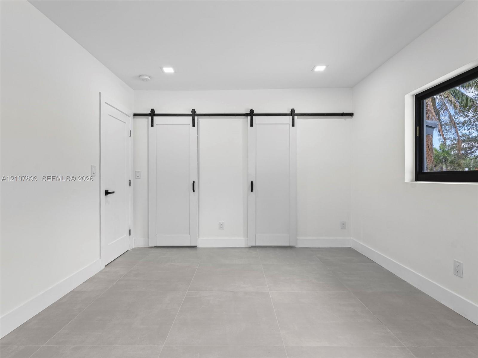 Interior room featuring large-format ceramic tile flooring, dual white sliding barn doors with matte black hardware, recessed ceiling lighting, and a window with a black frame