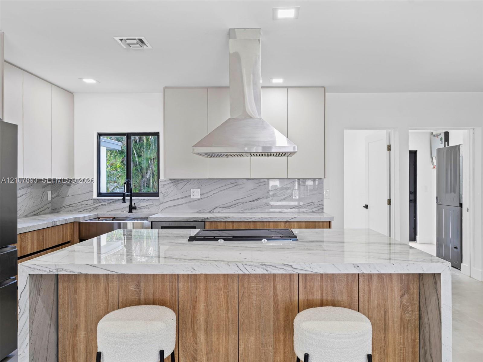 Kitchen featuring a waterfall-edge stone island with a cooktop, wood-finish cabinetry, stone slab backsplash, stainless steel chimney range hood, and matte black faucet