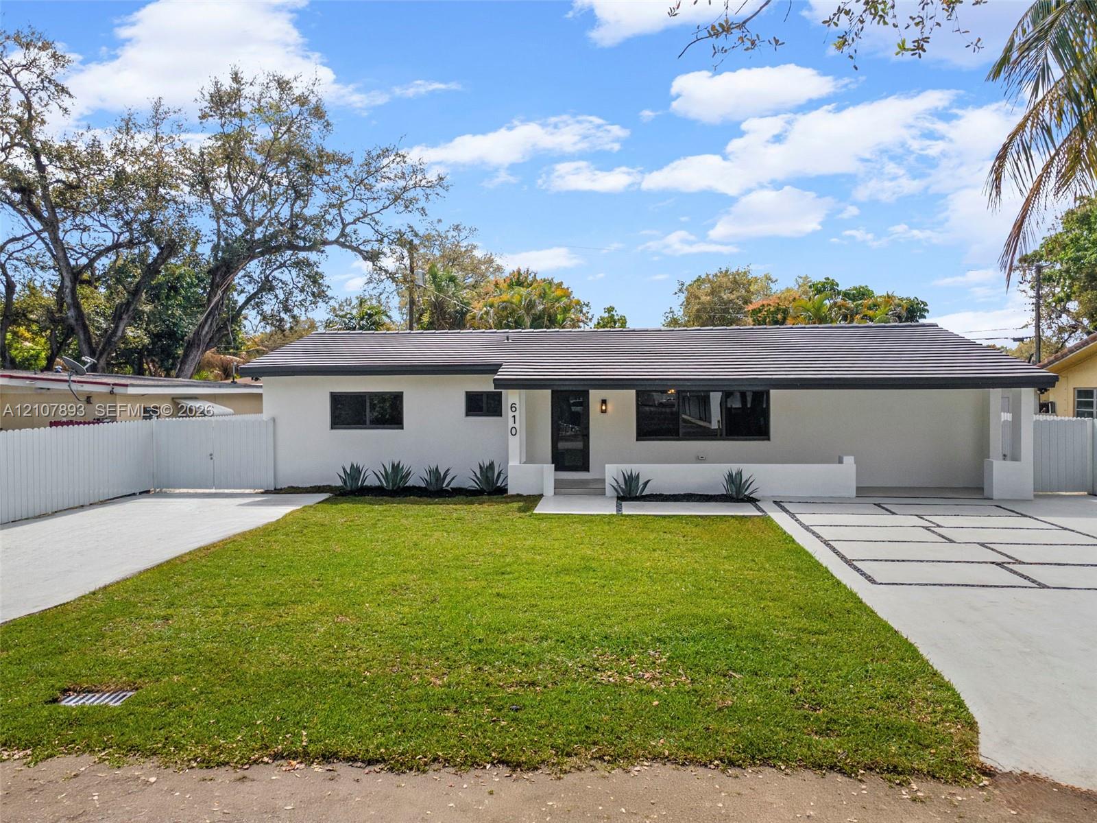 Single-story exterior featuring white stucco finish, dark-toned roof, paved geometric driveway, white perimeter fencing, and manicured lawn