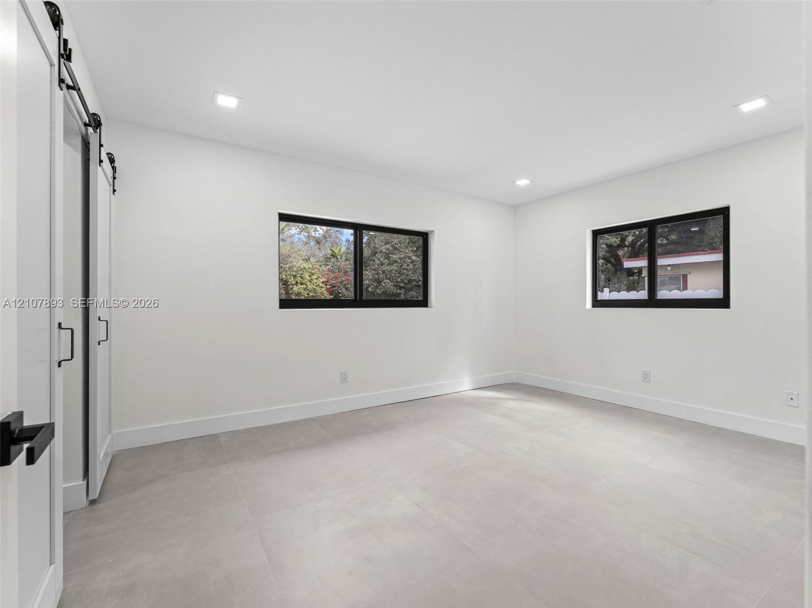 Interior space featuring neutral large-format tile flooring, recessed lighting, dual-pane windows with black frames, and a white sliding barn door with matte black hardware