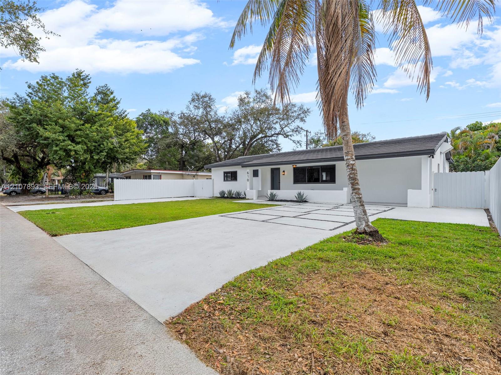 Single-story exterior featuring a painted stucco finish, attached carport, grid-patterned concrete driveway, white perimeter fencing, and established palm trees