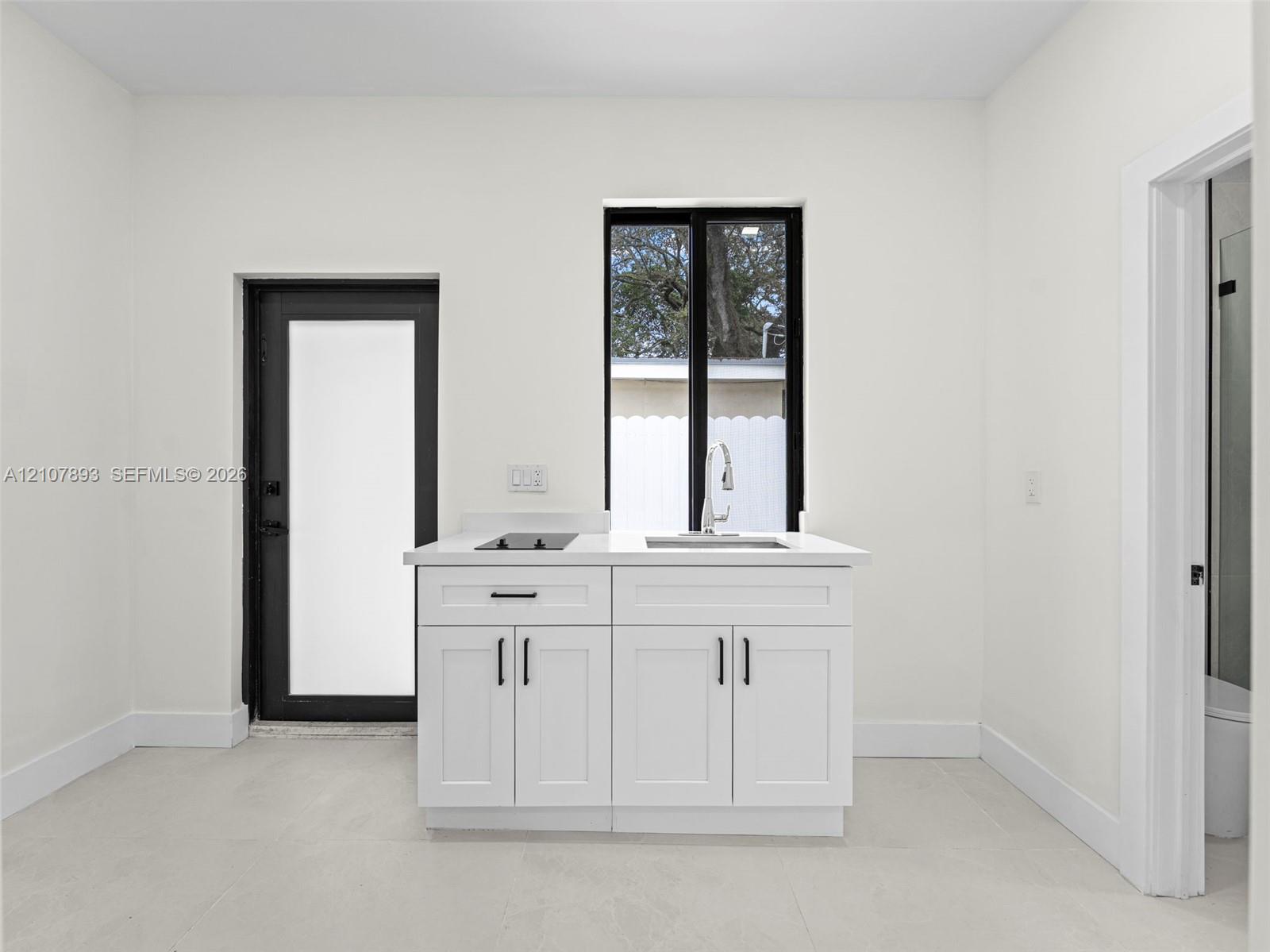Kitchenette featuring white shaker-style cabinetry, a light-toned countertop, a stainless steel sink with a high-arch faucet, a built-in two-burner induction cooktop, and contrasting black window and door frames