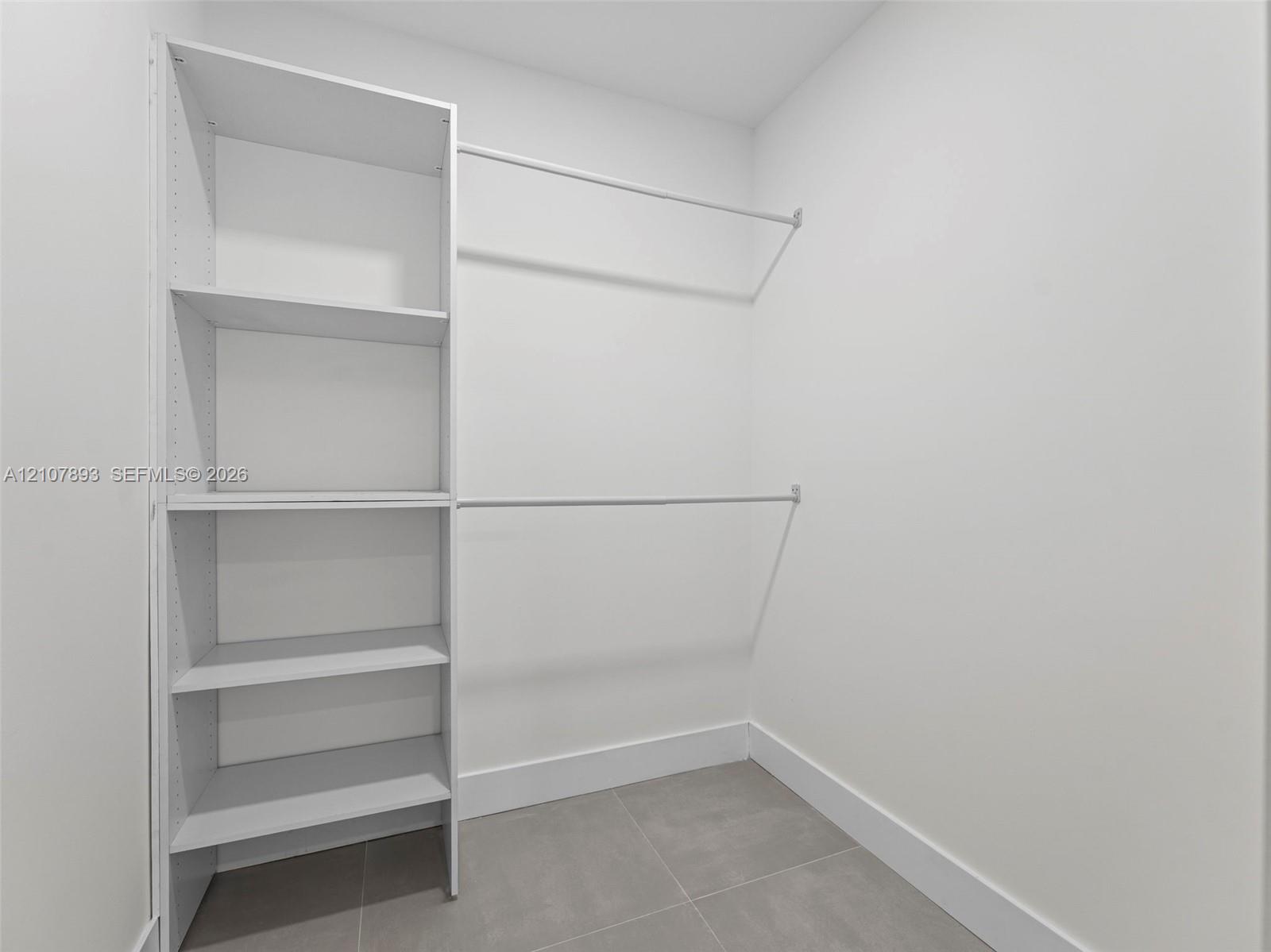Walk-in closet featuring white built-in shelving and dual-tier metal hanging rods with neutral tile flooring
