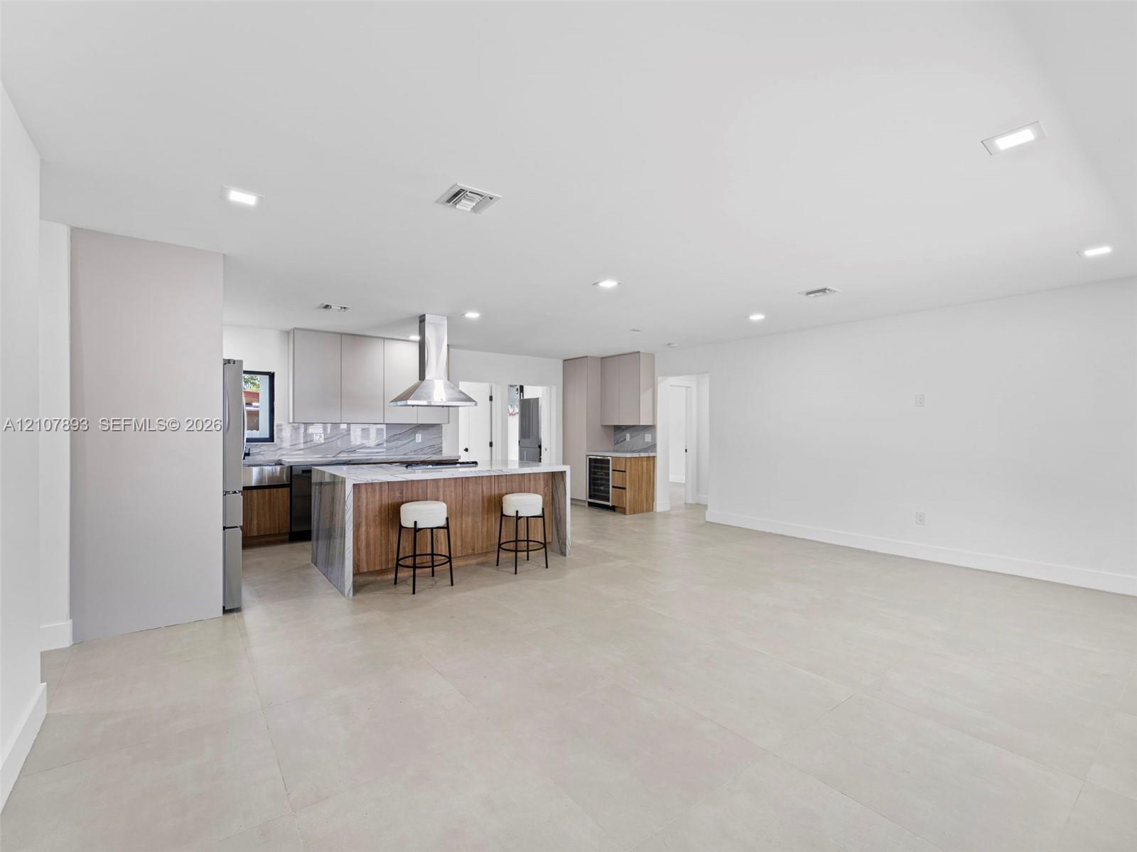 Open-concept kitchen featuring an oversized island with stone-finish waterfall edges, stainless steel range hood, built-in wine cooler, flat-panel cabinetry, and large-format tile flooring