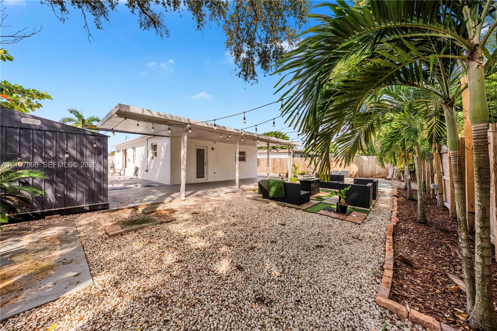 Rear exterior featuring a covered patio, gravel landscaping, perimeter wood fencing, and established palm trees
