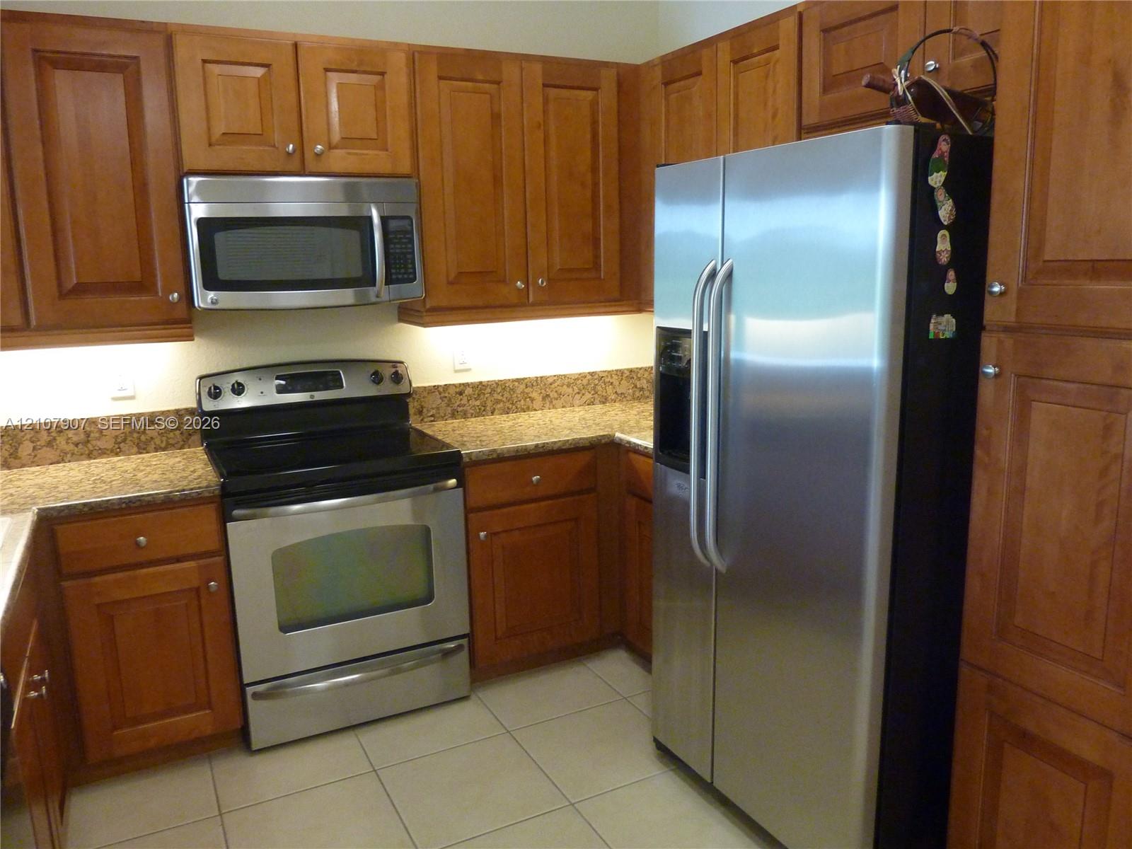 Kitchen featuring natural wood cabinetry, stone-surface countertops, tile flooring, and stainless steel appliances including a built-in microwave and side-by-side refrigerator