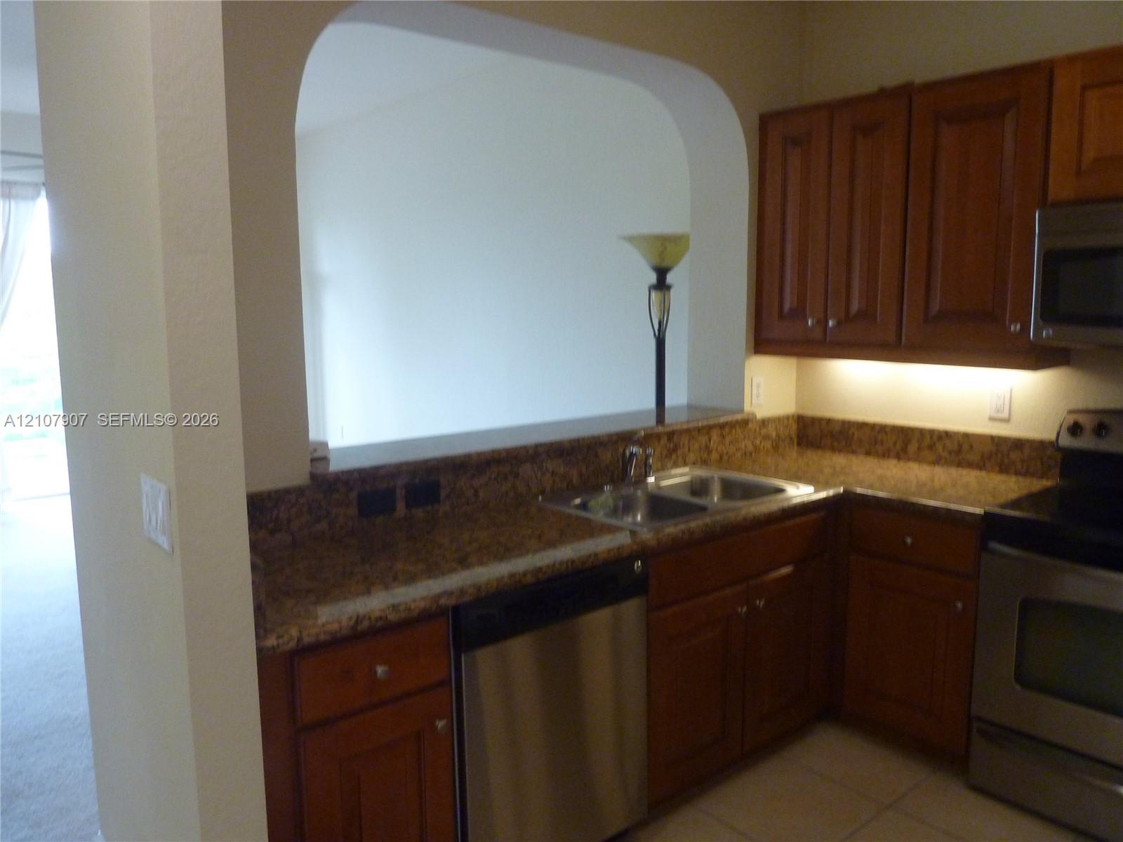 Kitchen featuring stone countertops, natural wood cabinetry, stainless steel appliances, a double-basin sink, and an arched pass-through window