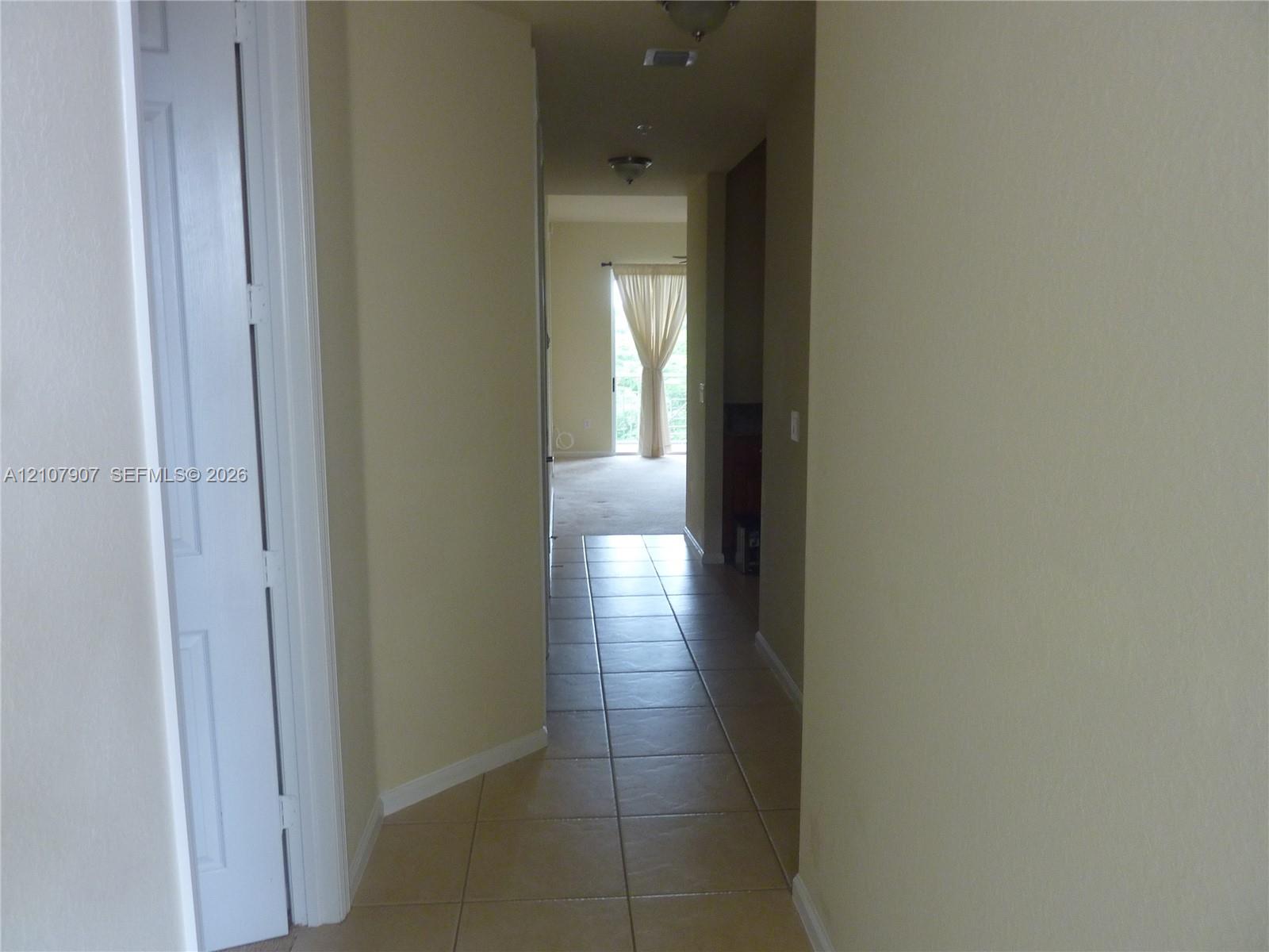 Interior hallway featuring neutral wall paint, tile flooring, two flush-mount ceiling light fixtures, and multi-panel interior doors