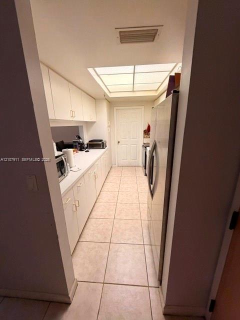 Galley kitchen featuring white cabinetry, light-toned tiled flooring, a skylight, and a stainless steel refrigerator