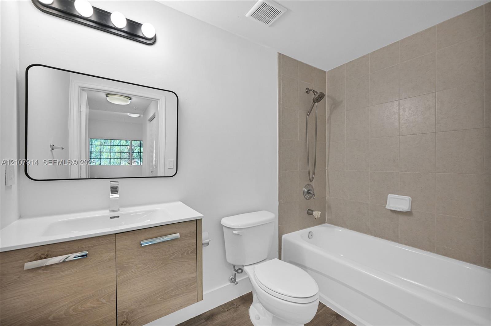 Floating wood-finish vanity with an integrated sink, rectangular mirror with a black frame, and a shower-tub combination featuring square tile surround and a handheld showerhead
