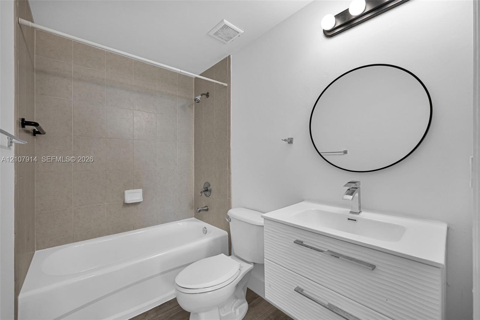 Modern bathroom featuring a floating vanity with a white countertop, circular wall-mounted mirror, three-light fixture, and bathtub with neutral wall tile