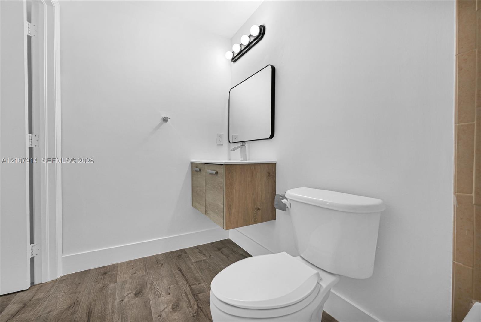 Floating wood-finish vanity with countertop, rectangular mirror with black frame, four-light vanity fixture, and wood-finish flooring