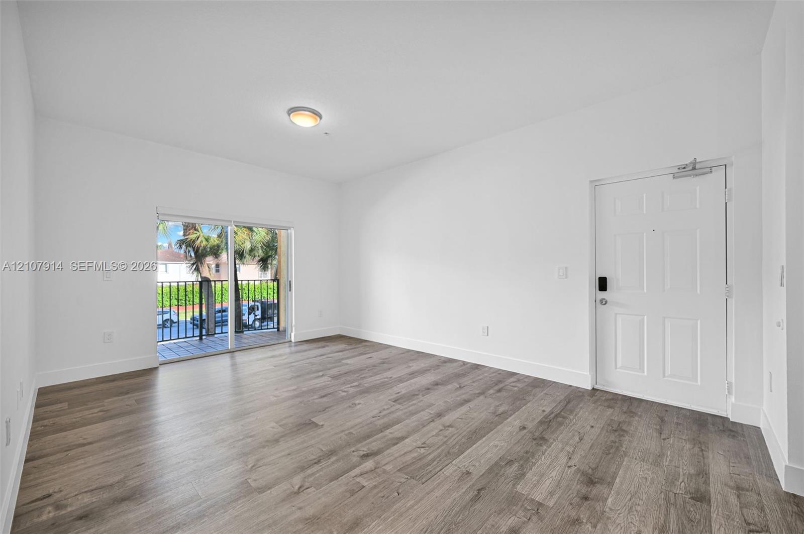 Open-concept interior featuring wood-finish flooring, white walls, flush-mount ceiling lighting, a multi-panel entry door, and sliding glass doors to a balcony with metal railing