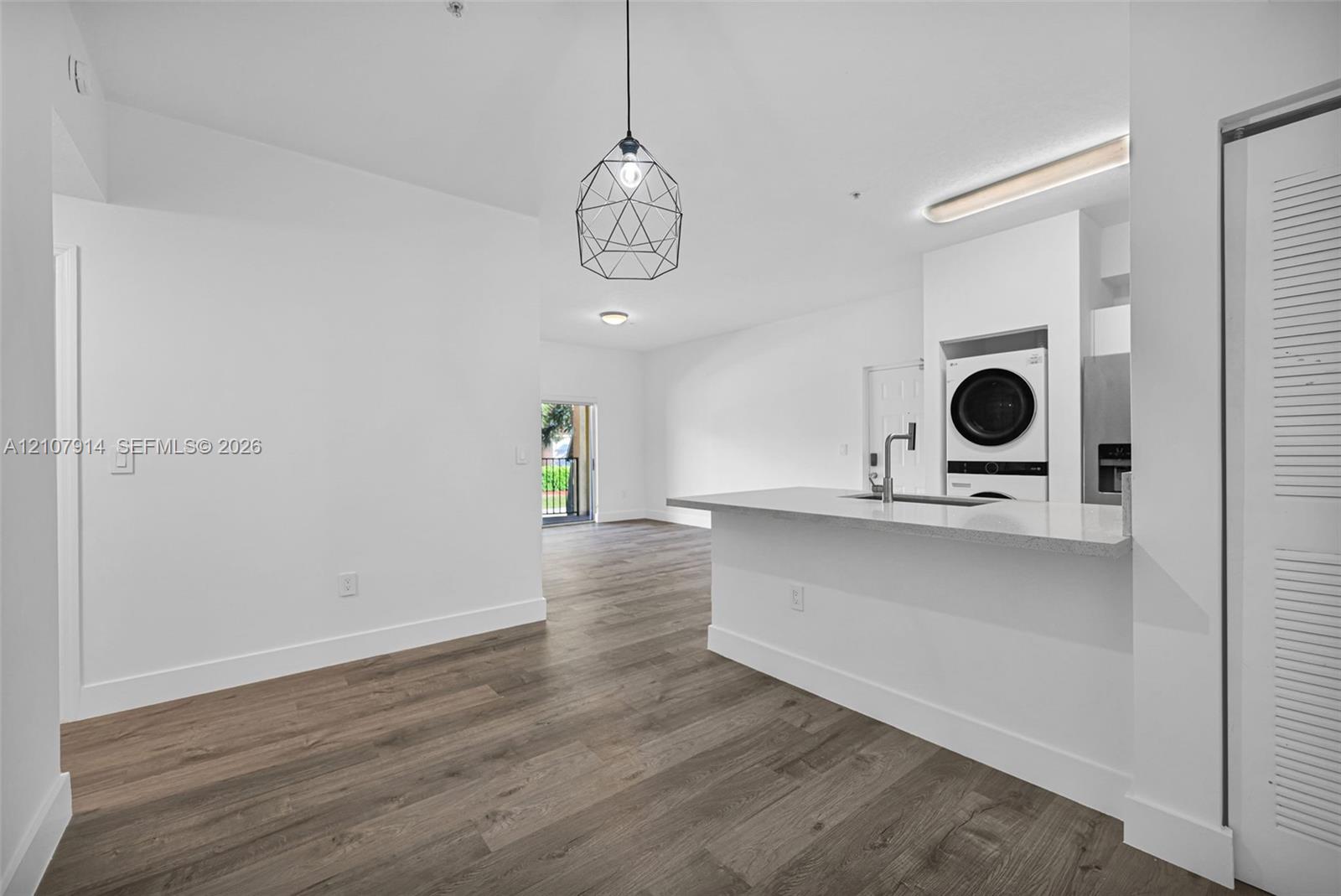 Kitchen featuring a quartz countertop island with an integrated sink, geometric pendant lighting, wood-finish flooring, and built-in stacked laundry appliances