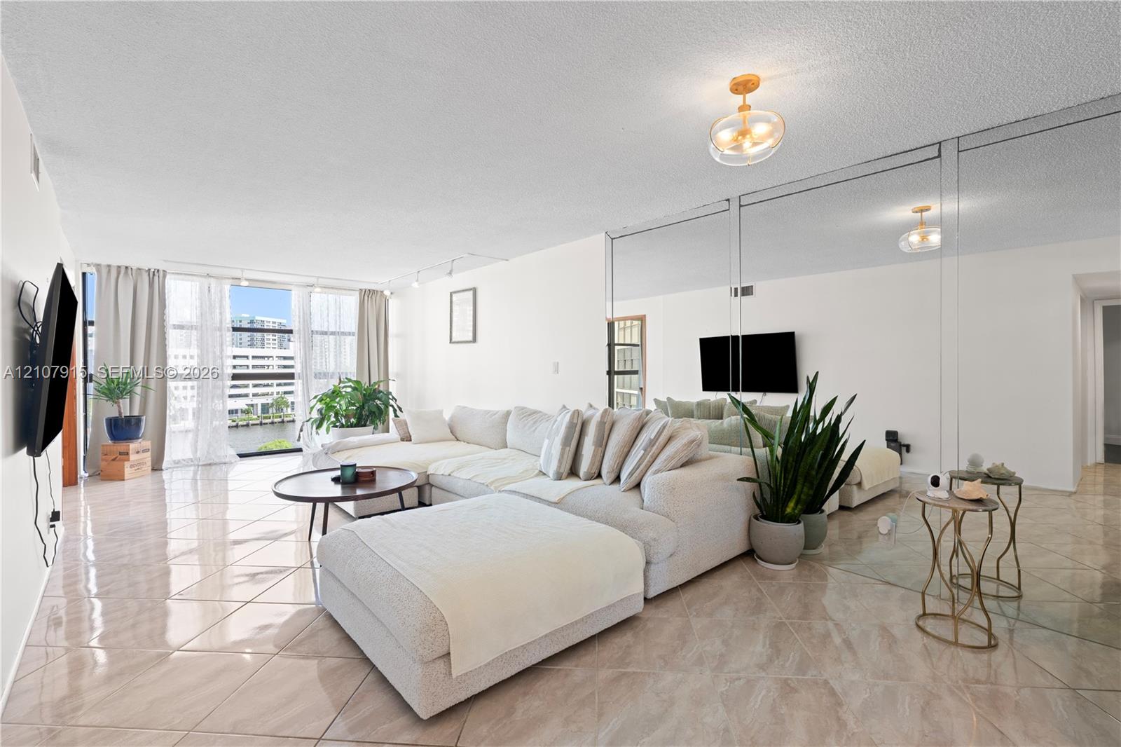 Living area featuring oversized floor-to-ceiling windows, polished tile flooring, full-wall mirrored paneling, and a contemporary flush-mount ceiling light fixture Living area featuring oversized floor-to-ceiling windows, polished tile flooring, full-wall mirrored paneling, and a contemporary flush-mount ceiling light fixture