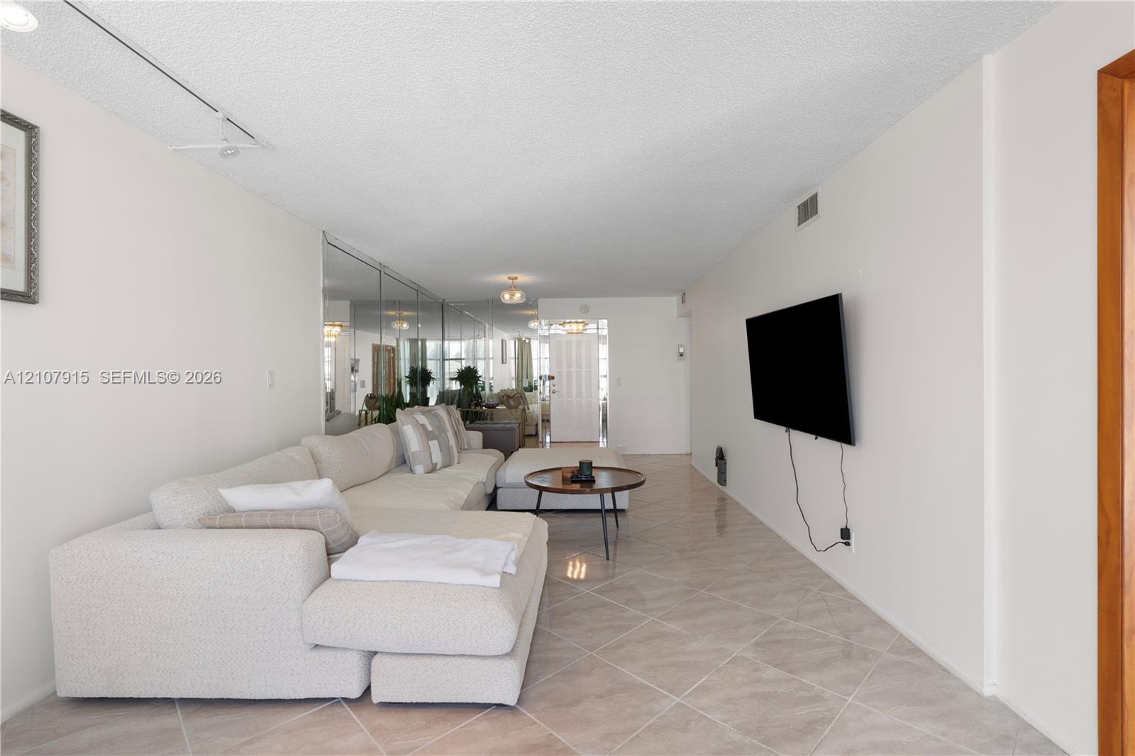 Spacious room featuring neutral tile flooring laid on a diagonal, full-height wall-mounted mirror paneling, textured ceiling, and track lighting Spacious room featuring neutral tile flooring laid on a diagonal, full-height wall-mounted mirror paneling, textured ceiling, and track lighting