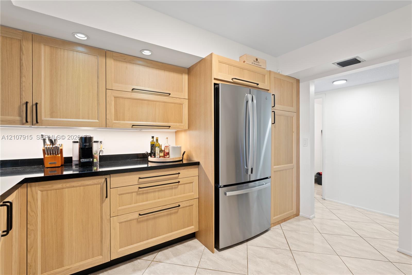 Light wood-finish cabinetry, dark stone countertops, stainless steel French-door refrigerator, under-cabinet lighting, and neutral ceramic tile flooring